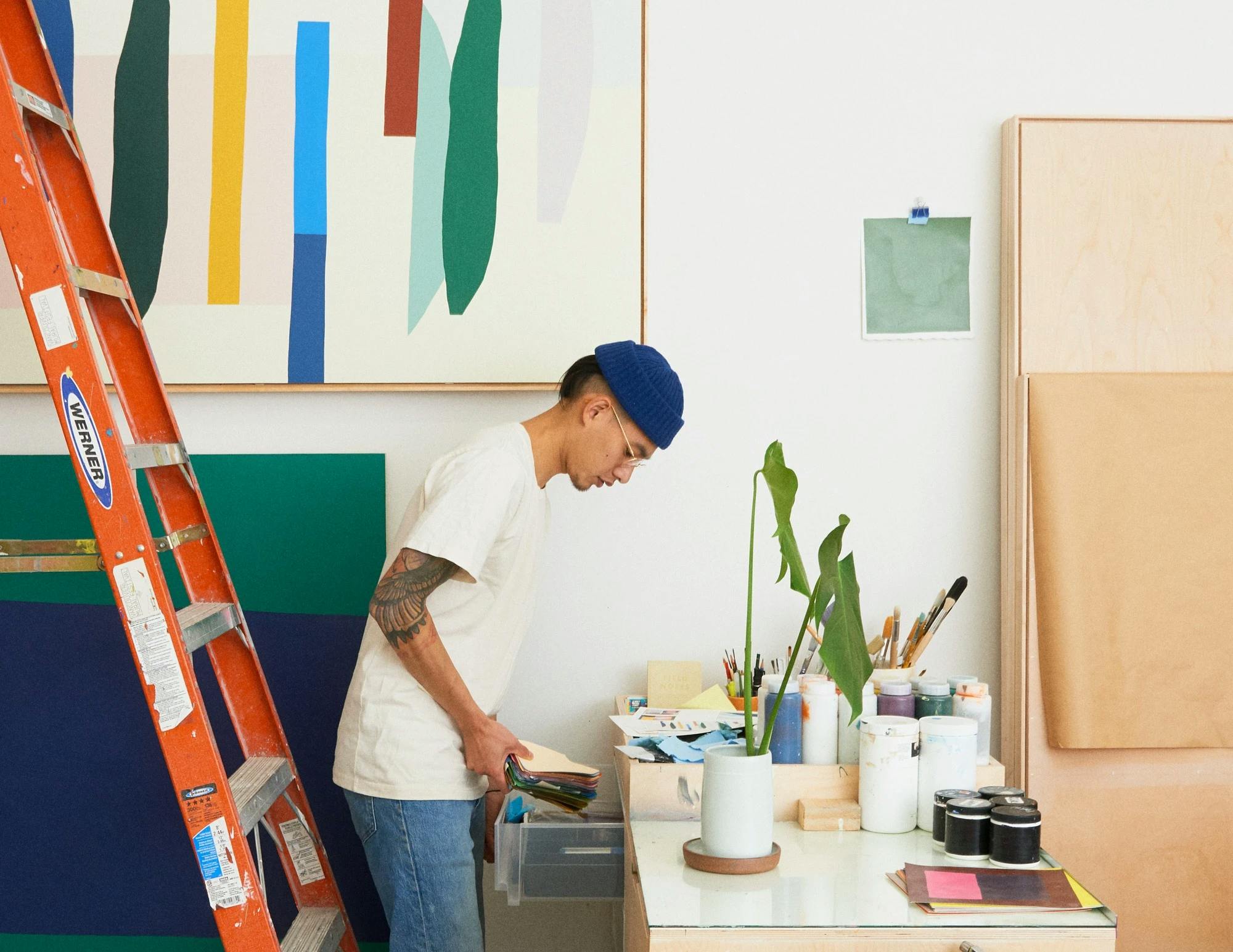 Artist Scott Sueme wearing a blue beanie hat, white t-shirt, and jeans working in his studio standing next to a red ladder.