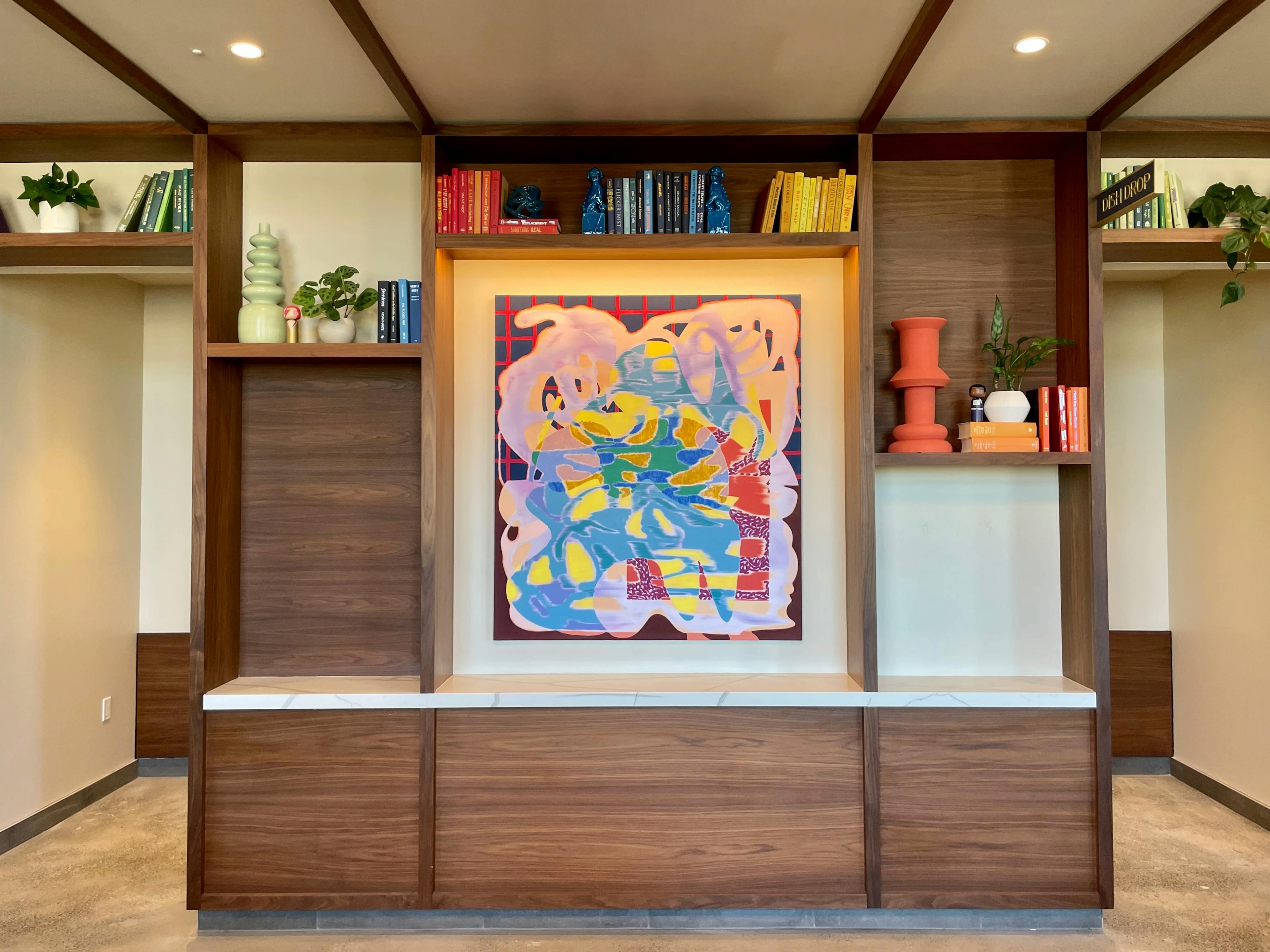 A large multicolored painting by artist Trina Turturici installed within wooden shelving next to books and sculptures.