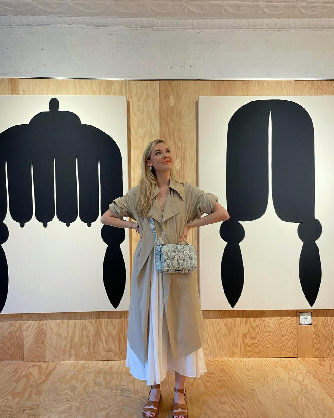 Woman in trench coat stands with hands on hips in wood-clad exhibition space showcasing abstract hairdo artworks at Uprise Art.