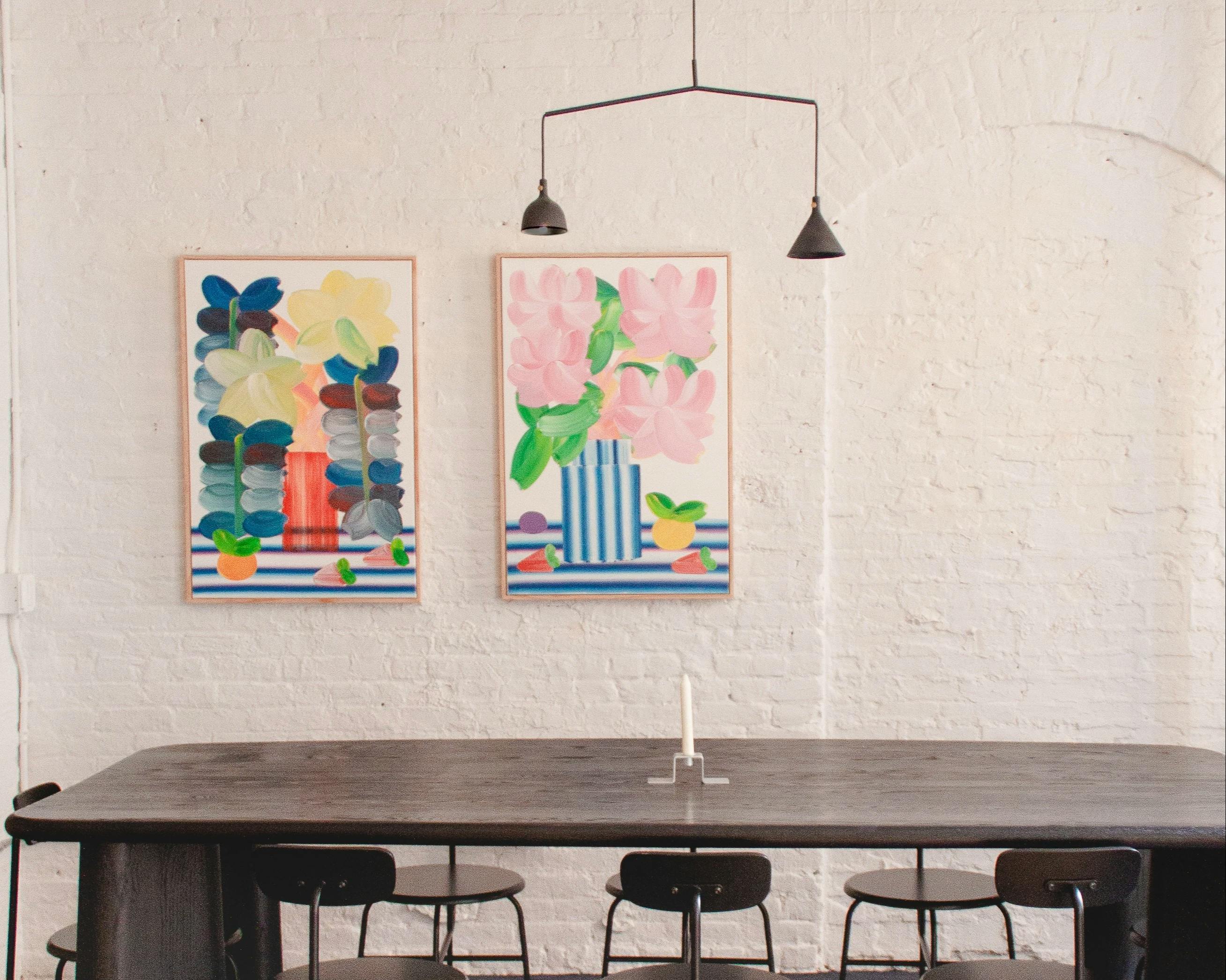 Two colorful still life paintings by artist Erin D. Garcia on a white brick wall above a black dining room table at Uprise Art.