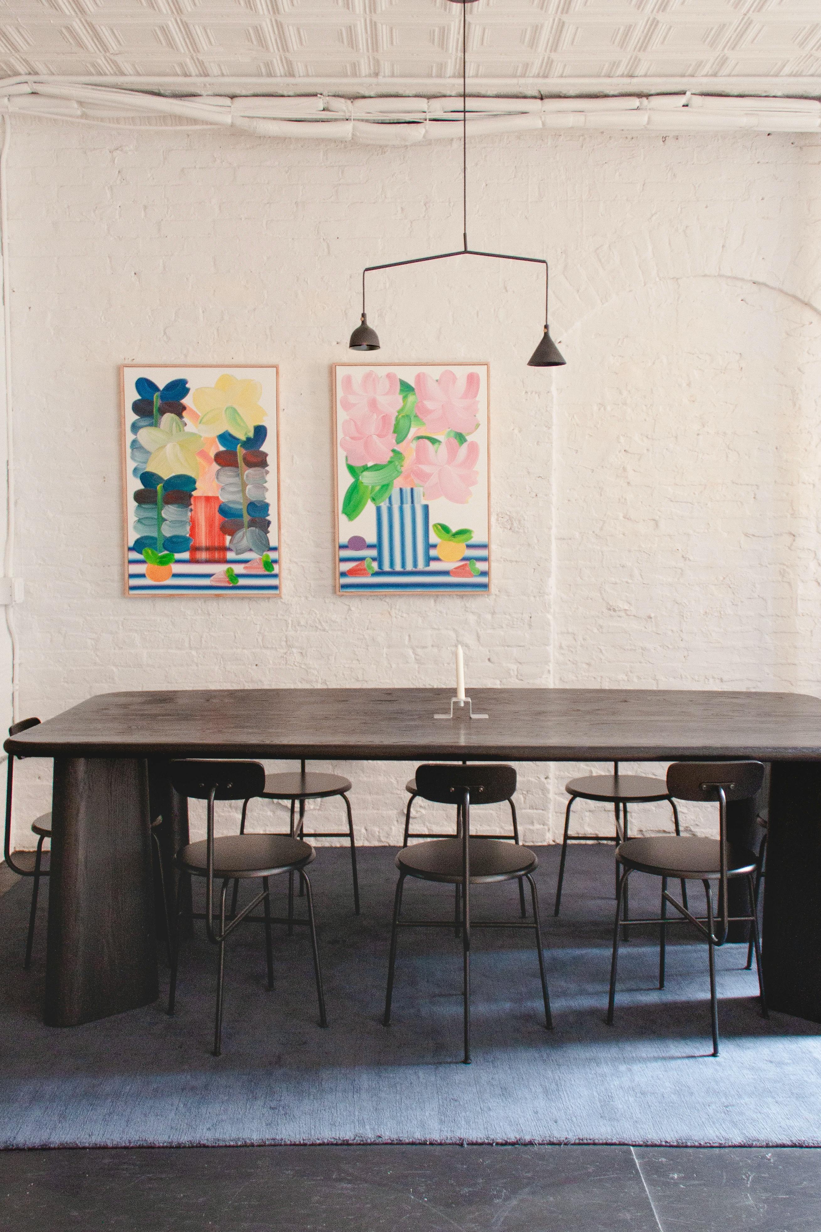 Two colorful still life paintings by artist Erin D. Garcia on a white brick wall above a black dining room table at Uprise Art.
