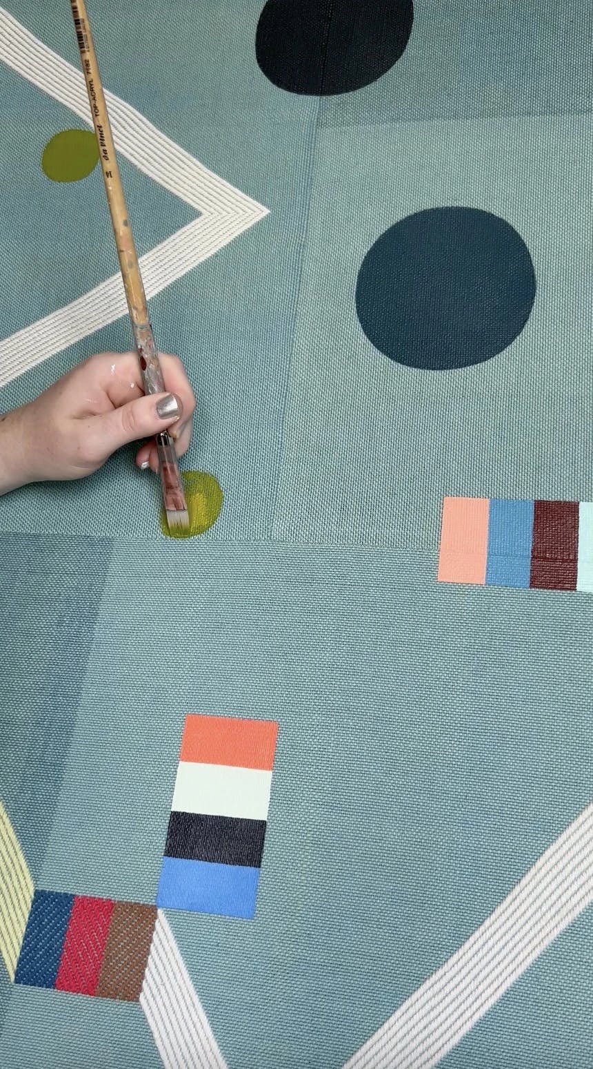 A close-up of artist Sarah Sullivan Sherrod holding a paintbrush and painting a small green circle on a handwoven canvas.