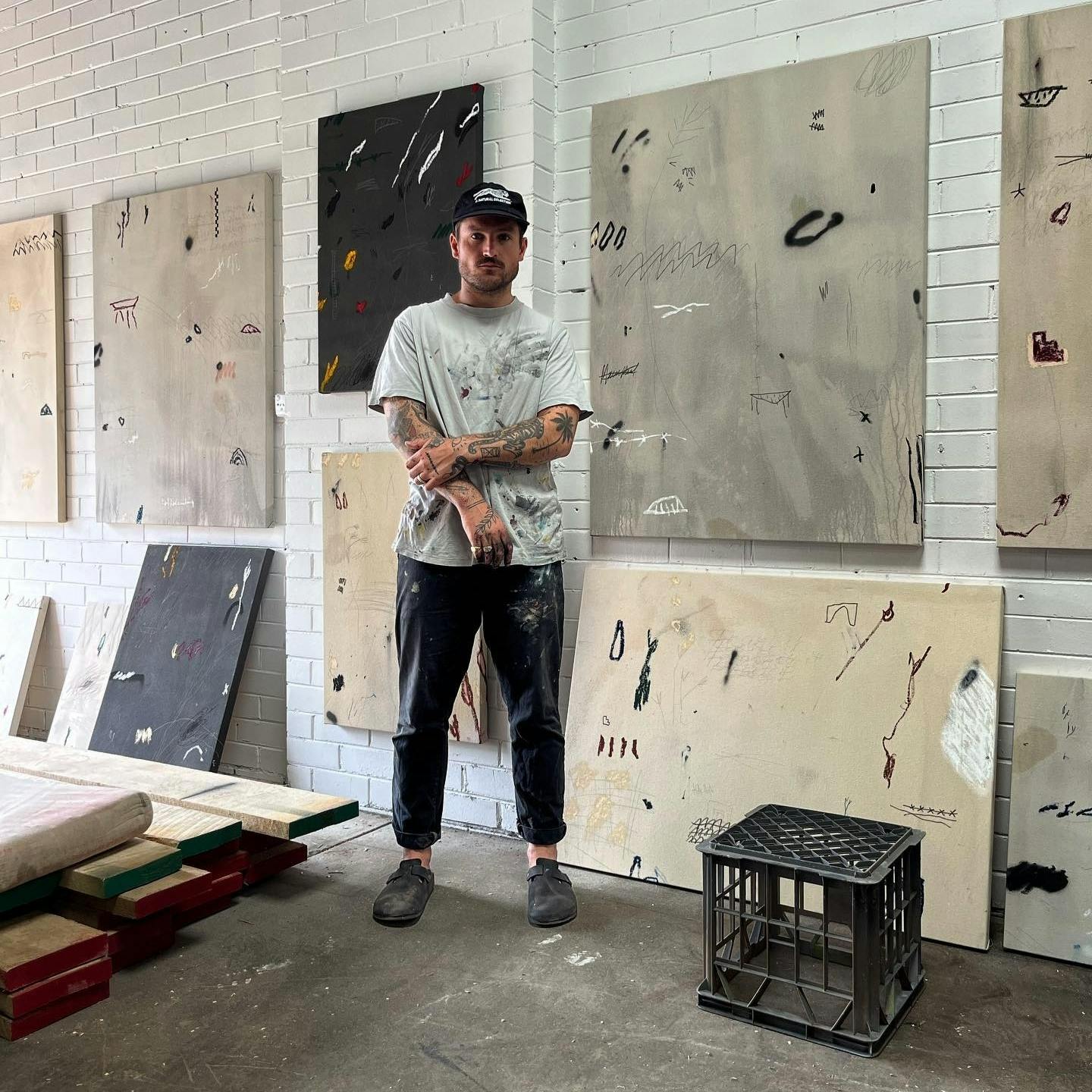 Artist Saxon Quinn standing in his studio with grey, tan, and black paintings hanging on the wall behind him.