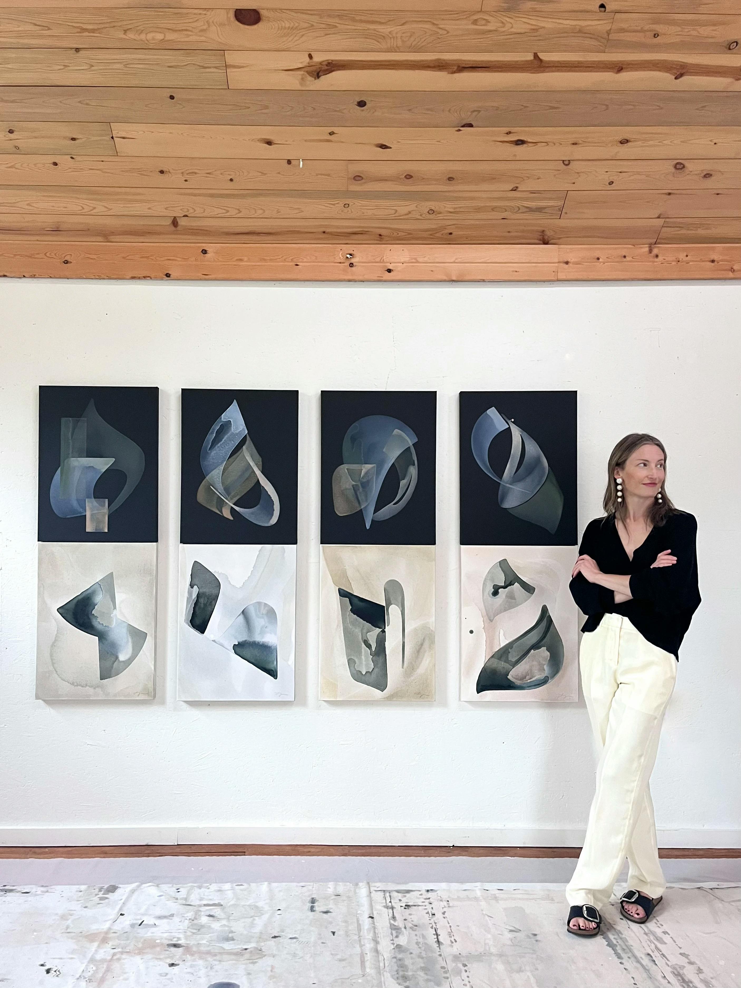 Artist Laura Naples standing in front of a grid of her black and beige acrylic wash paintings installed on a white wall in her studio.