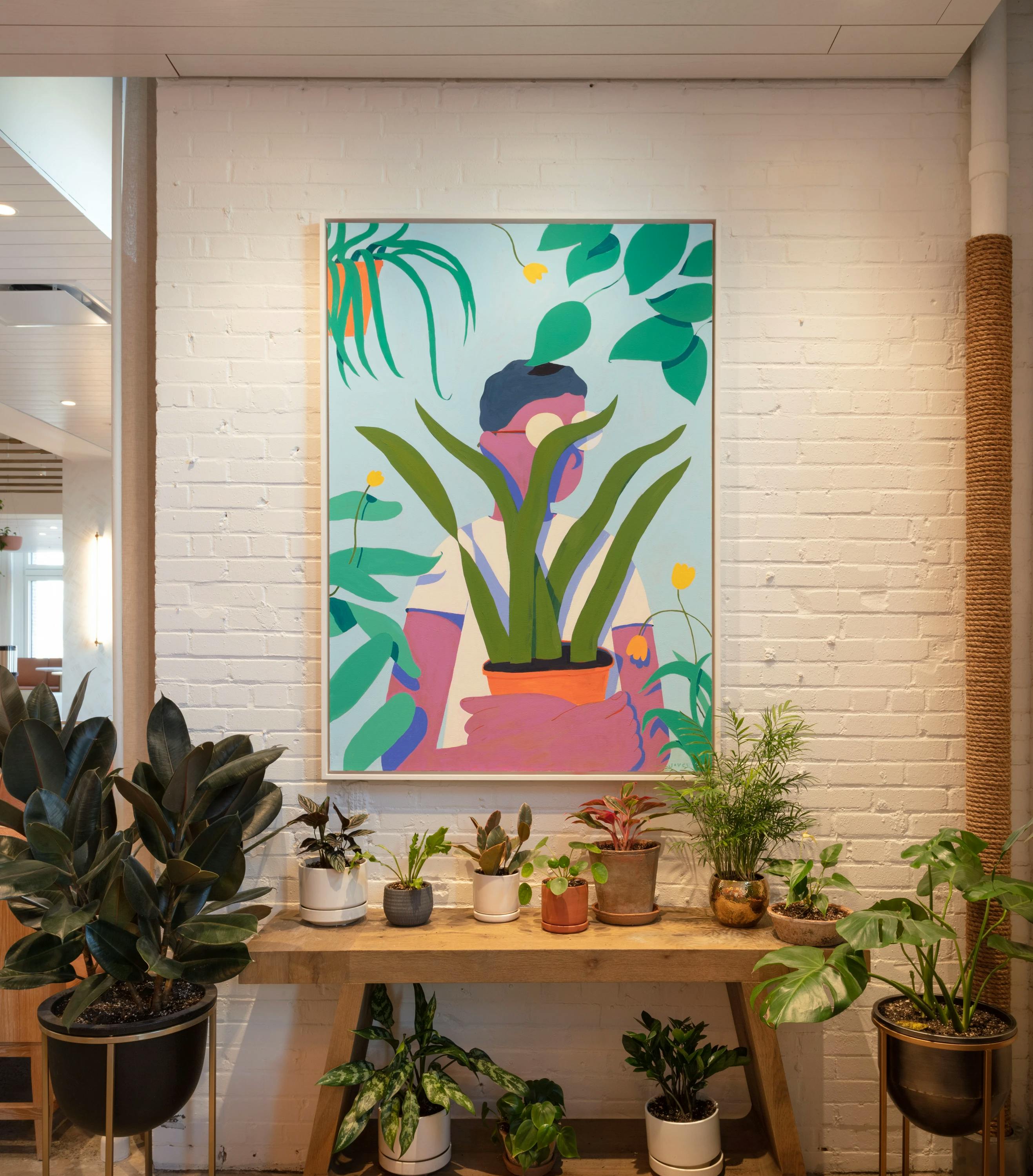 A painting by artist Jackson Joyce, depicting a person wearing glasses and standing among plants, installed above a table full of houseplants.