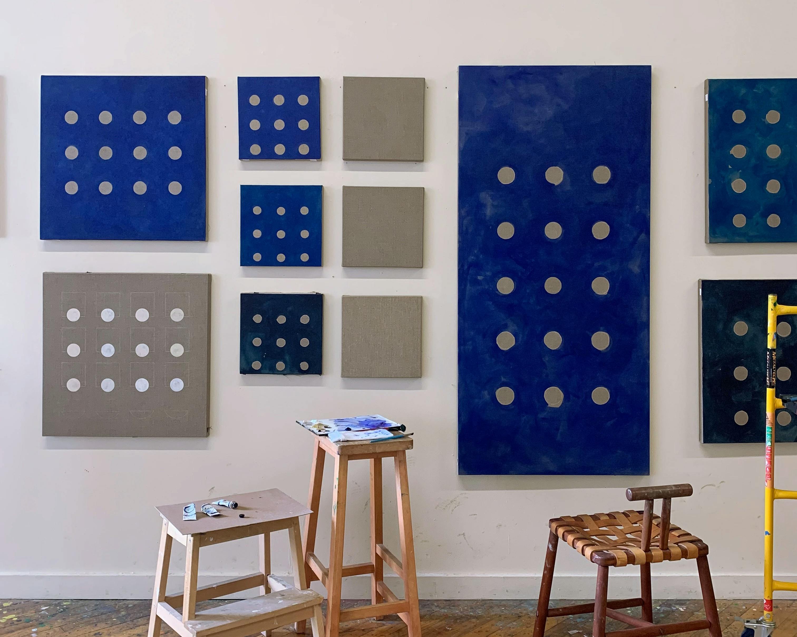 Minimalist, blue and grey paintings with grids of circles by artist Carla Weeks installed on a white wall in her studio.