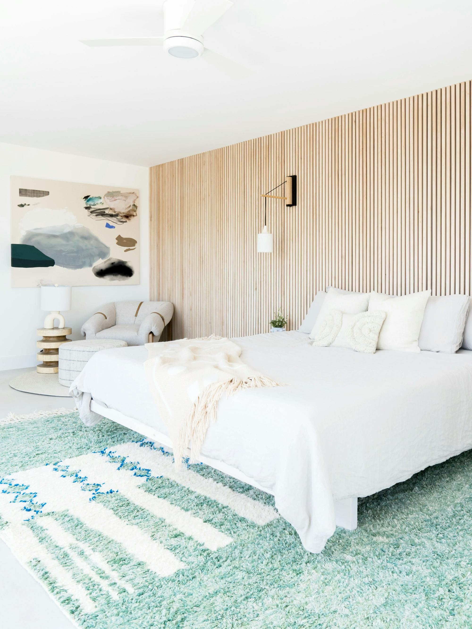 A neutral bedroom with a teal carpet, bed with white sheets, and an abstract painting by artist Karina Bania above a cushioned reading chair in the corner.