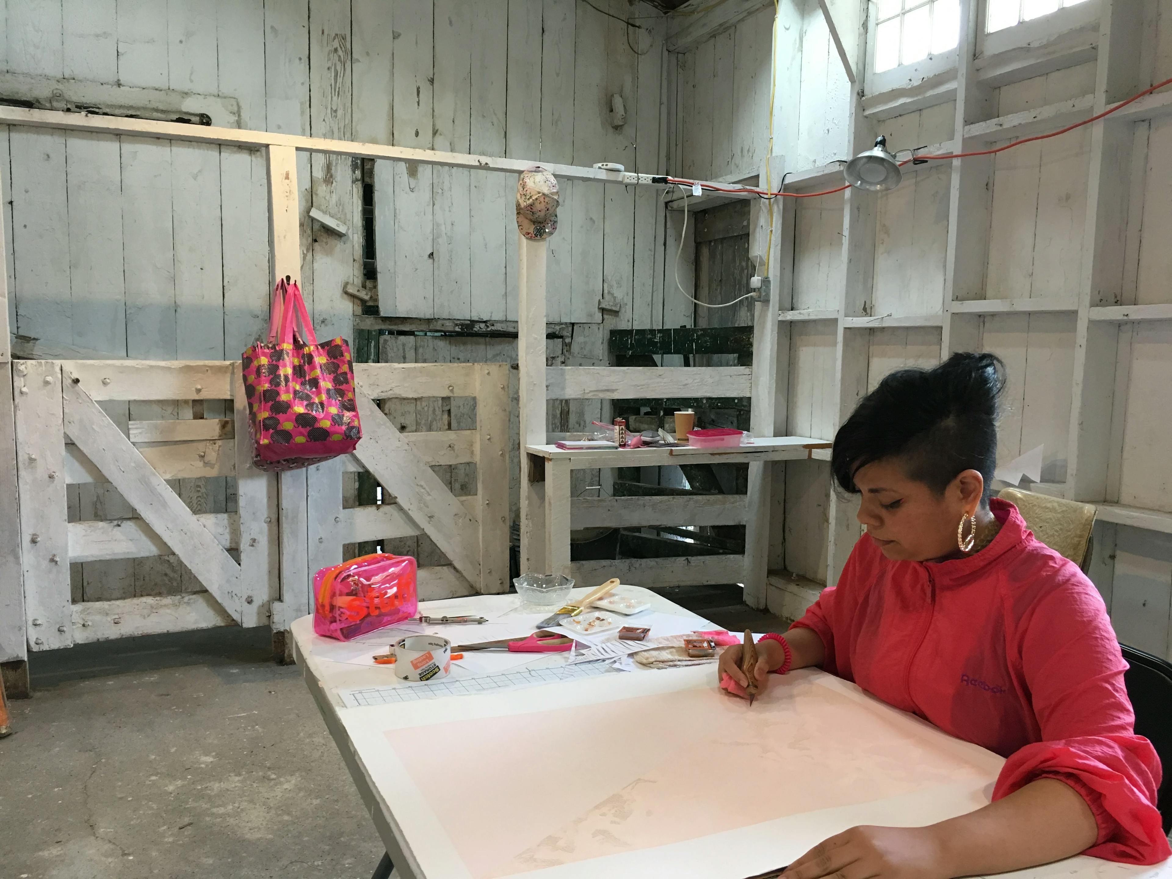 Artist Lucha Rodríguez sitting at a table in her studio, using a knife on her paper compositions.