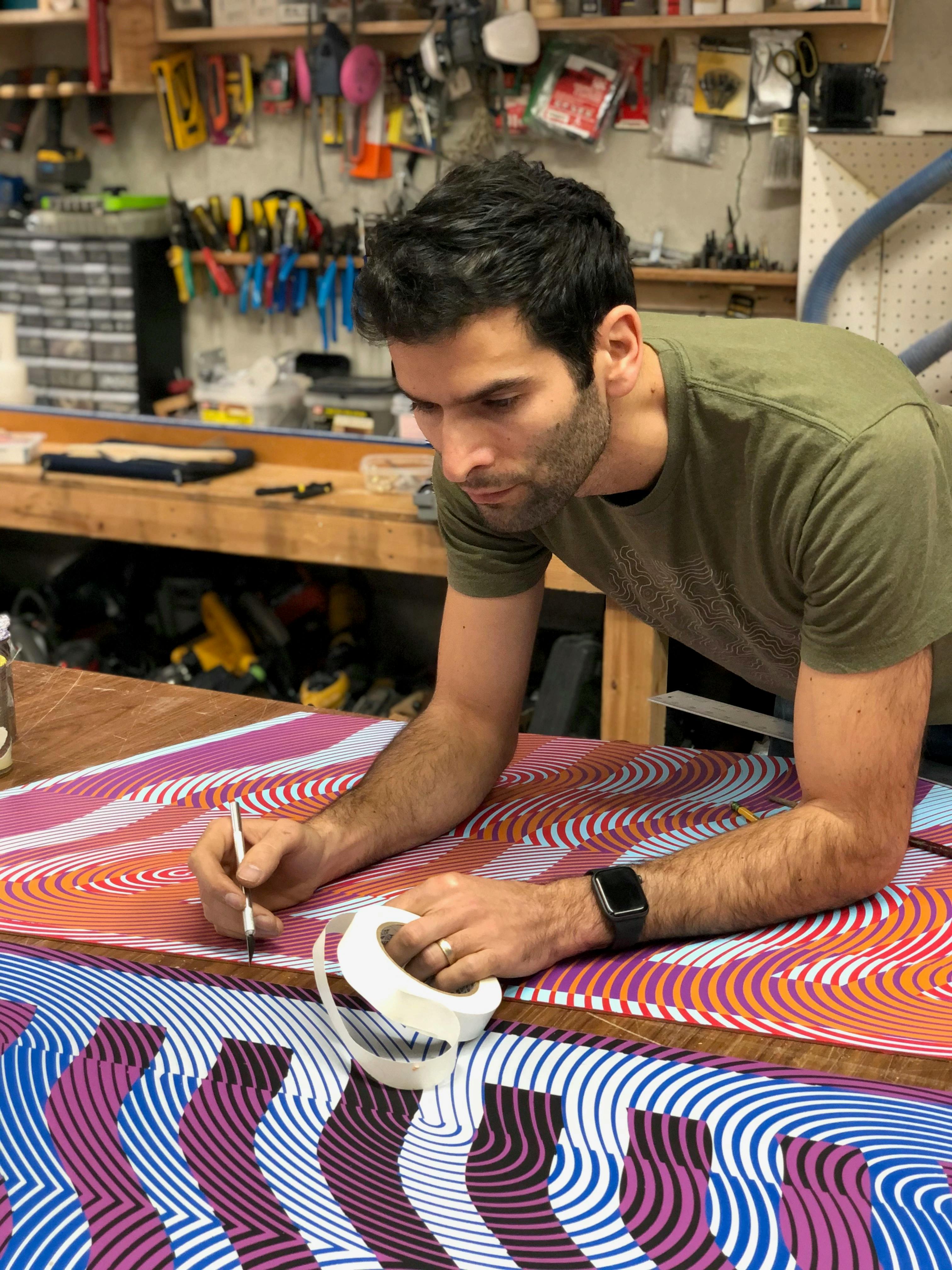 Artist Matt Neuman holding masking tape and leaning over abstract, colorful prints in his studio.