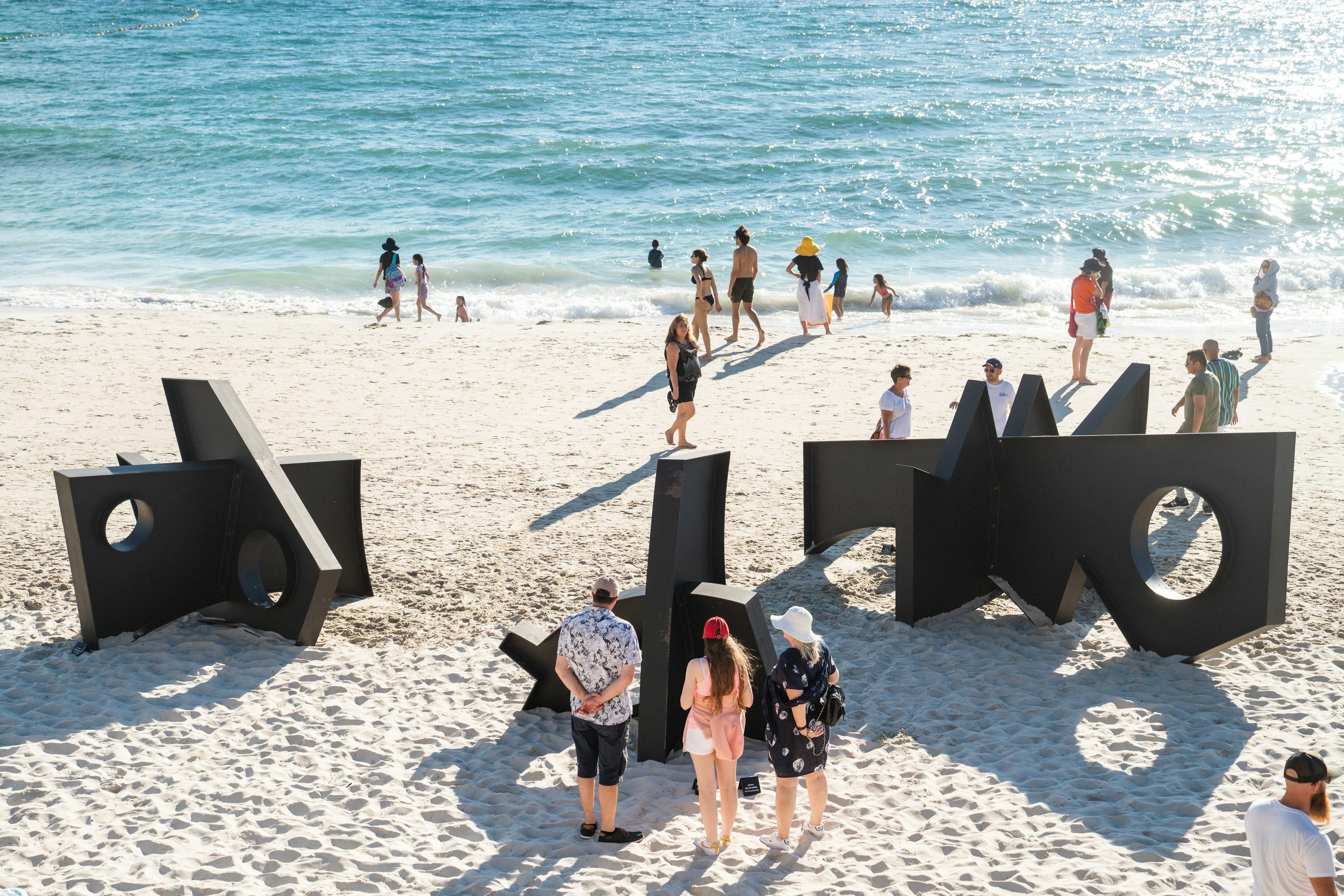 People walking around a public installation of three black metal sculptures by Fitzhugh Karol on a beach in Australia.