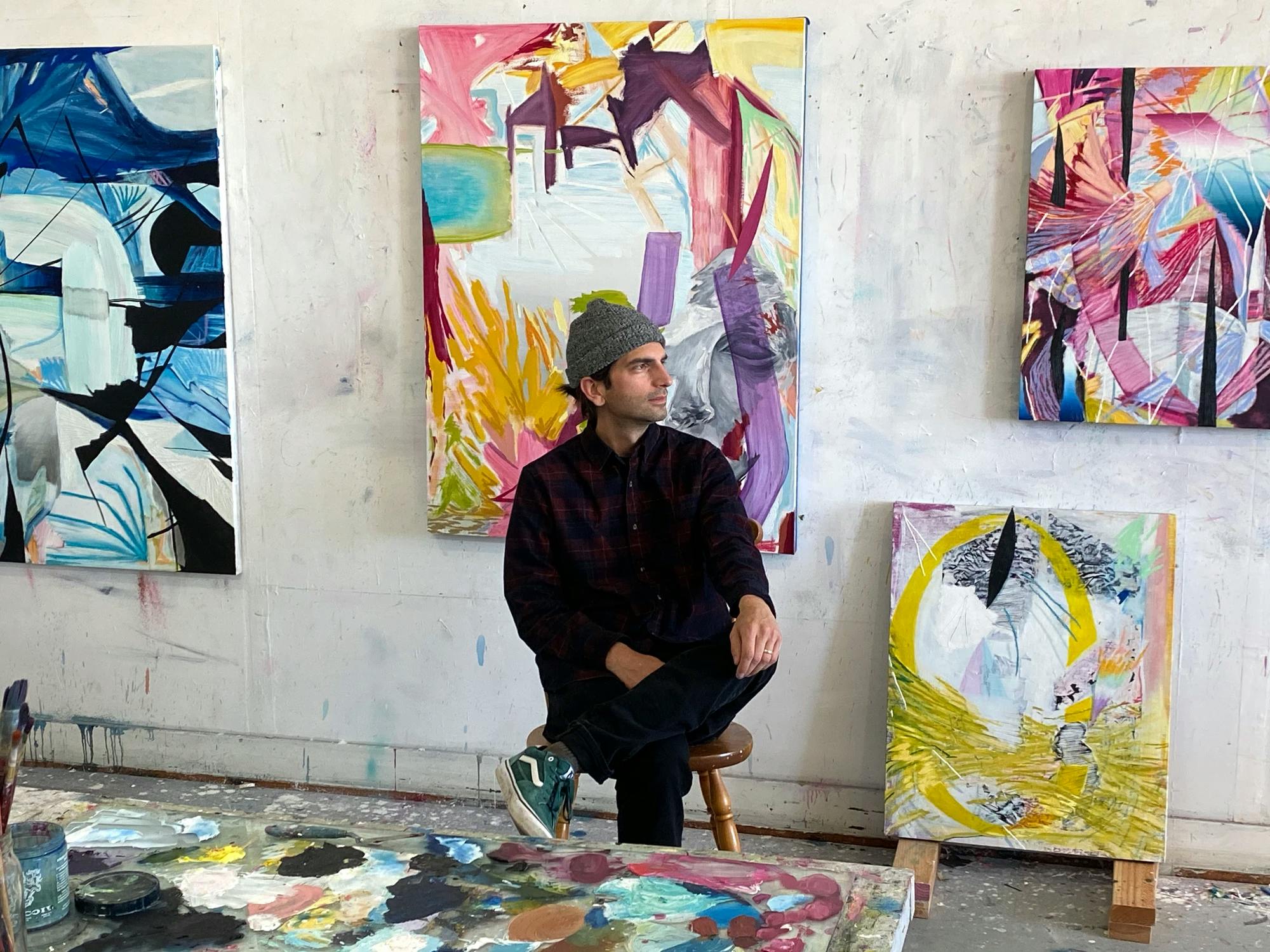 Artist Tyler Scheidt sitting on a stool in his studio surrounded by his abstract, colorful paintings.