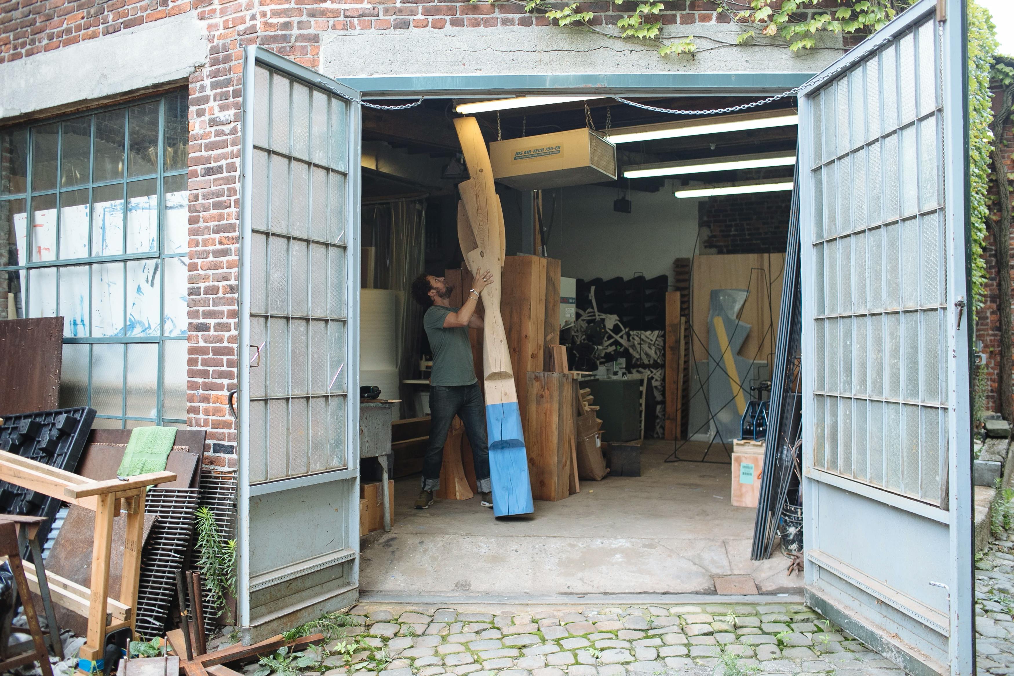 Artist Fitzhugh Karol holding a tall blue and beige wooden sculpture inside his double-doored studio.