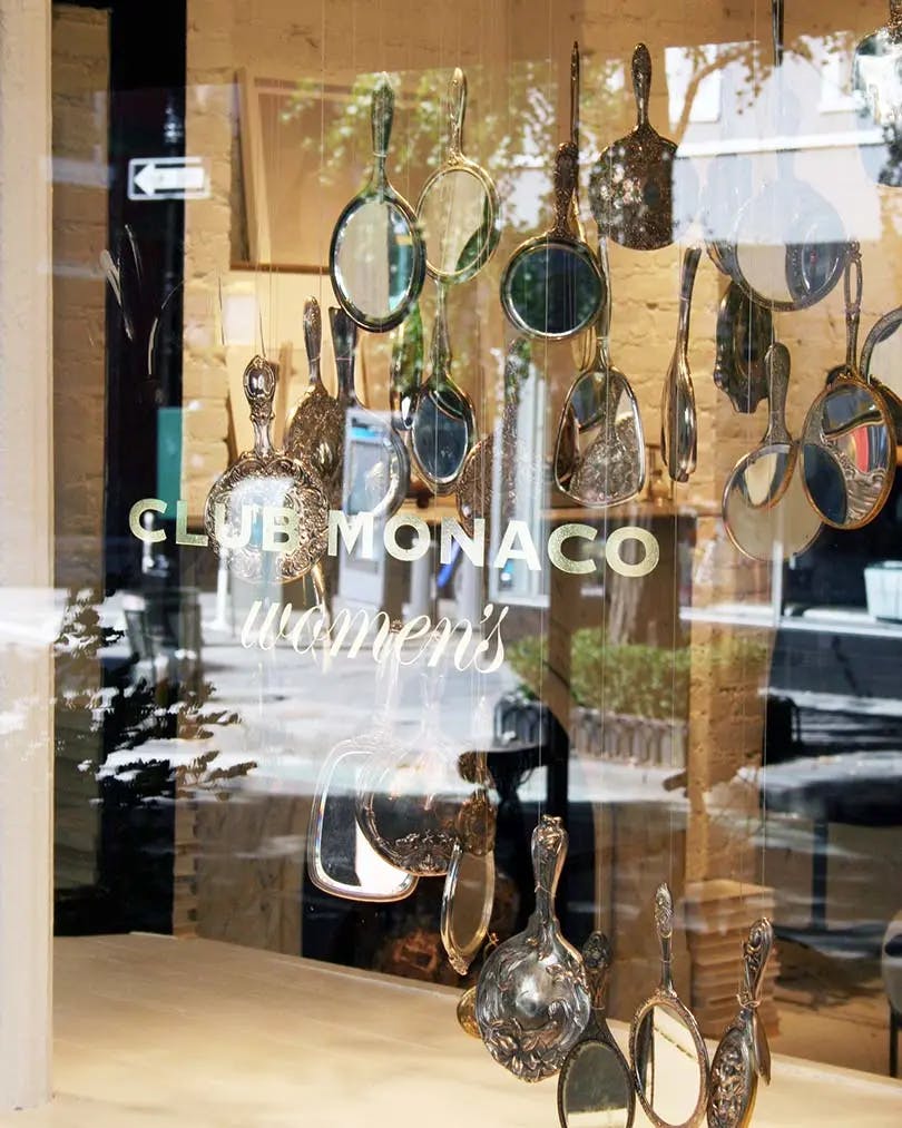 The window storefront etched with "Club Monaco women's" containing an installation of suspended hand-held mirrors inside. 