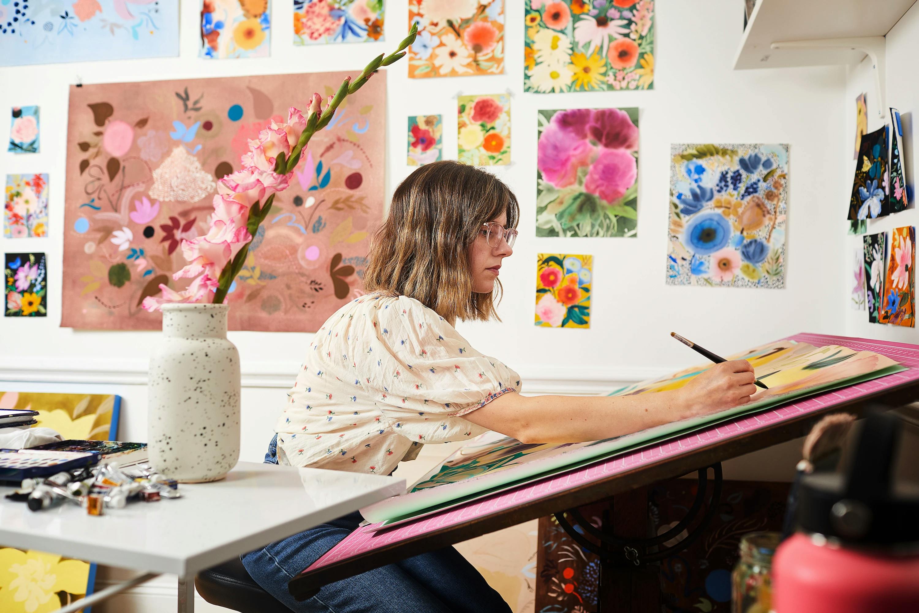 Artist Kayla Plosz Antiel sitting at a table in her studio working on a watercolor painting. She is surrounded by her floral artworks hanging on the walls around her.