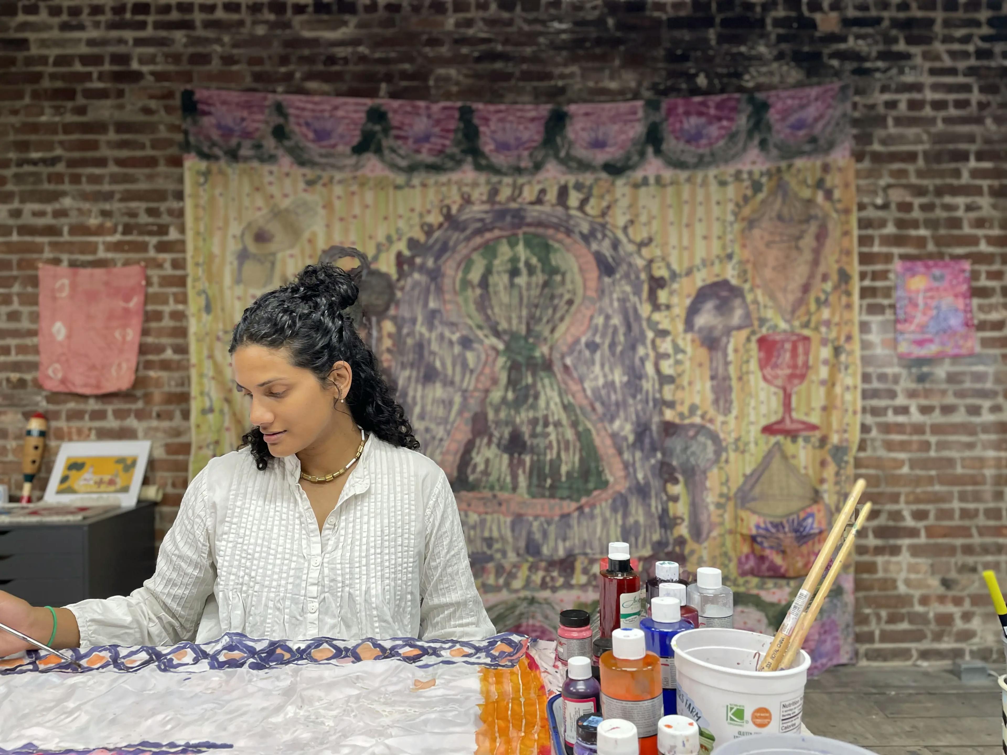 The artist Padma Rajendran working in her studio. The artist sits at a table painting on silk while one of her completed large scale silk paintings hangs behind her.