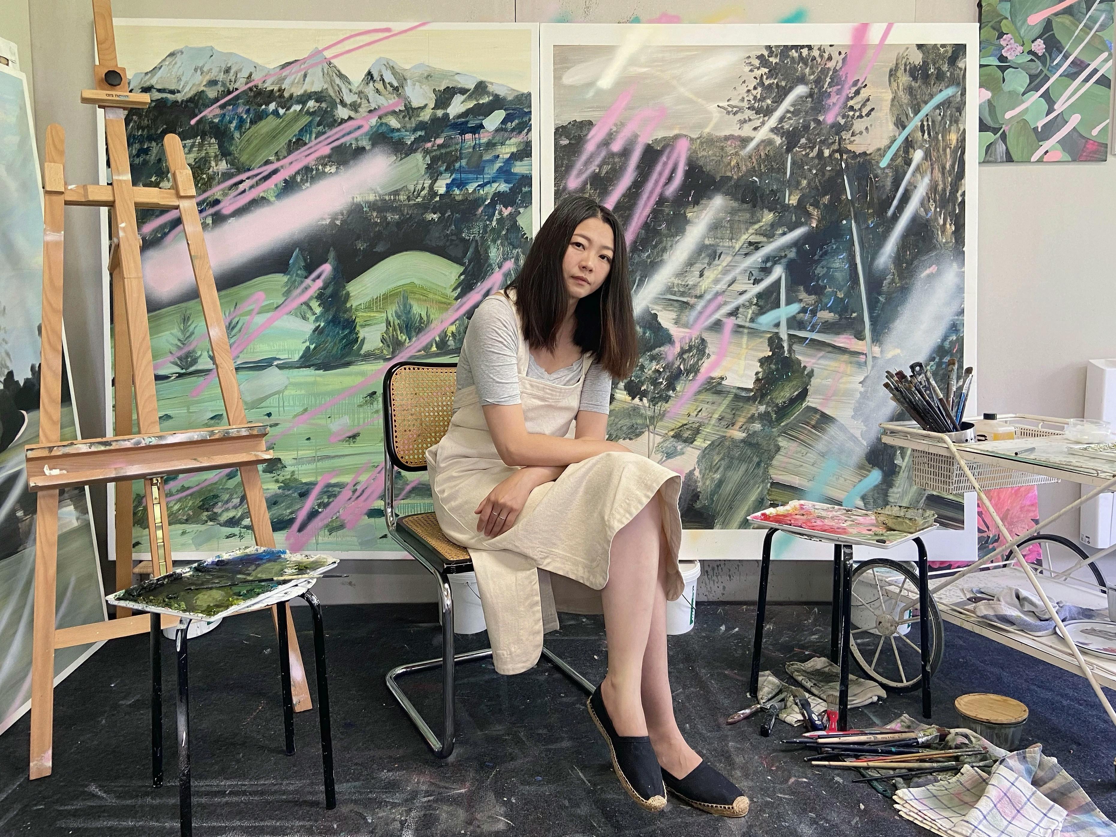 The artist Una Ursprung sitting in her studio in front of two large paintings that depict landscapes interspersed with abstract pink, and white spraypaint marks.