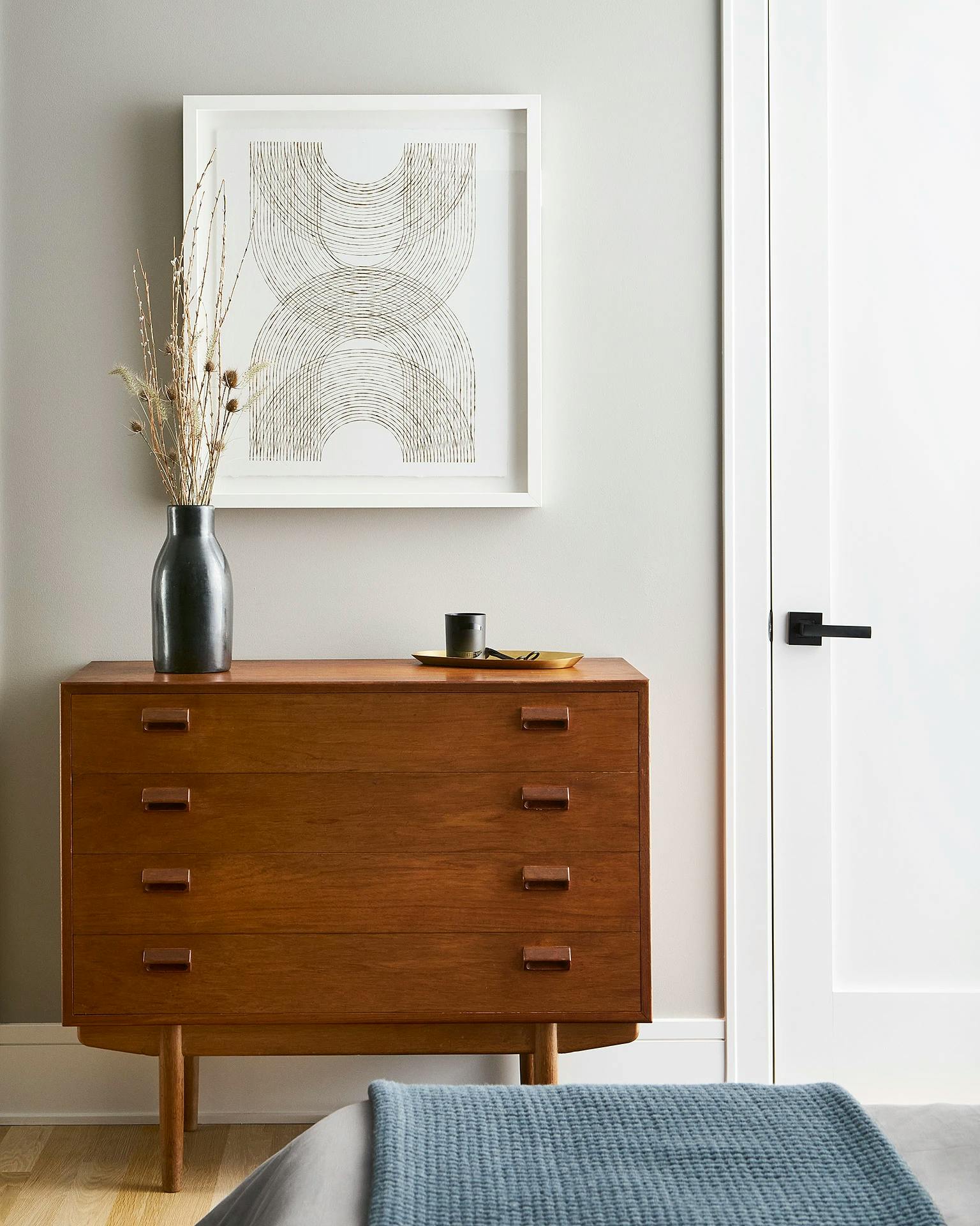 Framed geometric artwork with overlapping semicircles by artist Katrine Hildebrandt-Hussey installed on a white wall above a wooden dresser in a bedroom.
