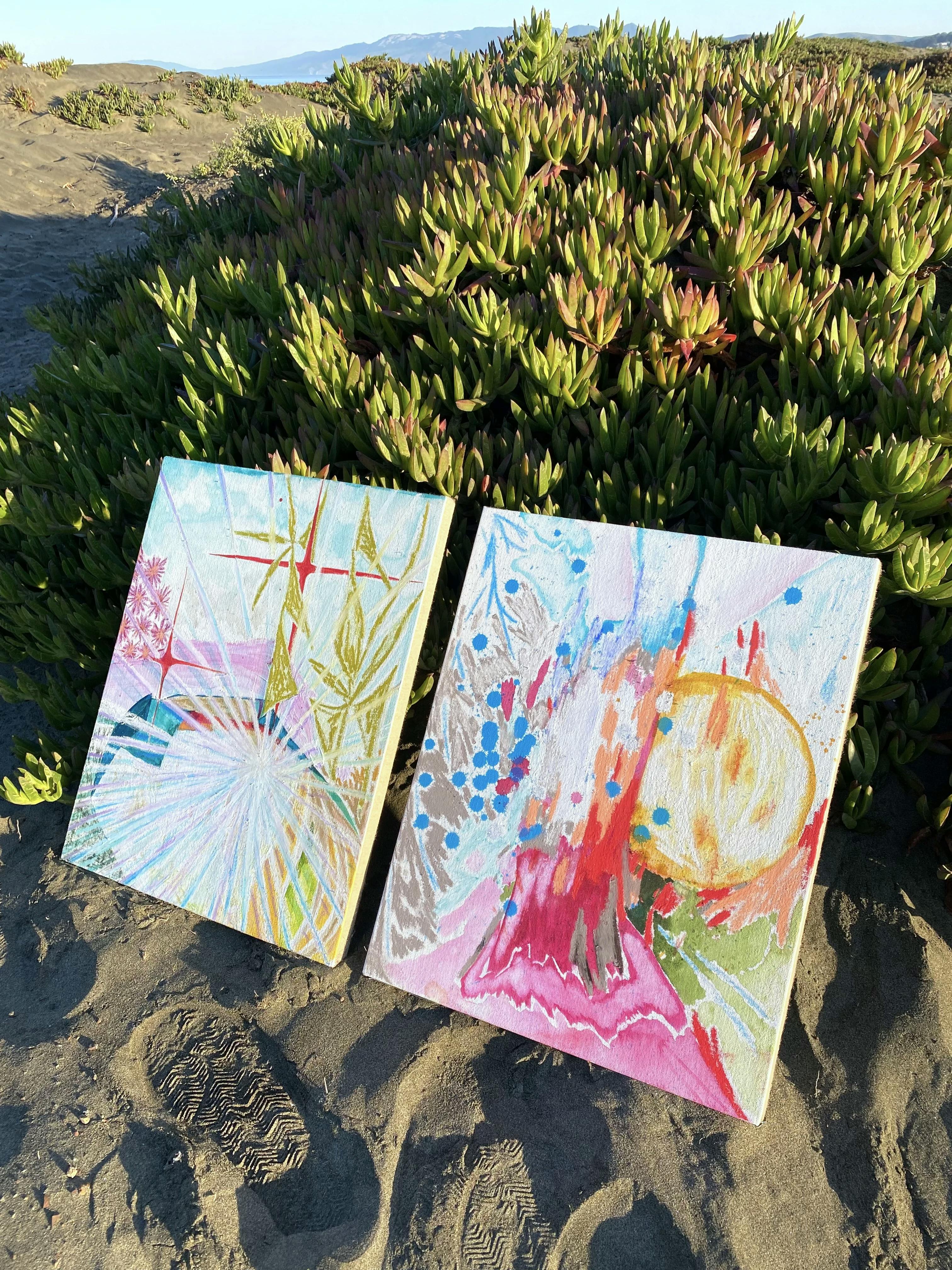 Two abstract paintings by artist Tyler Scheidt leaning against a bush on the sand.