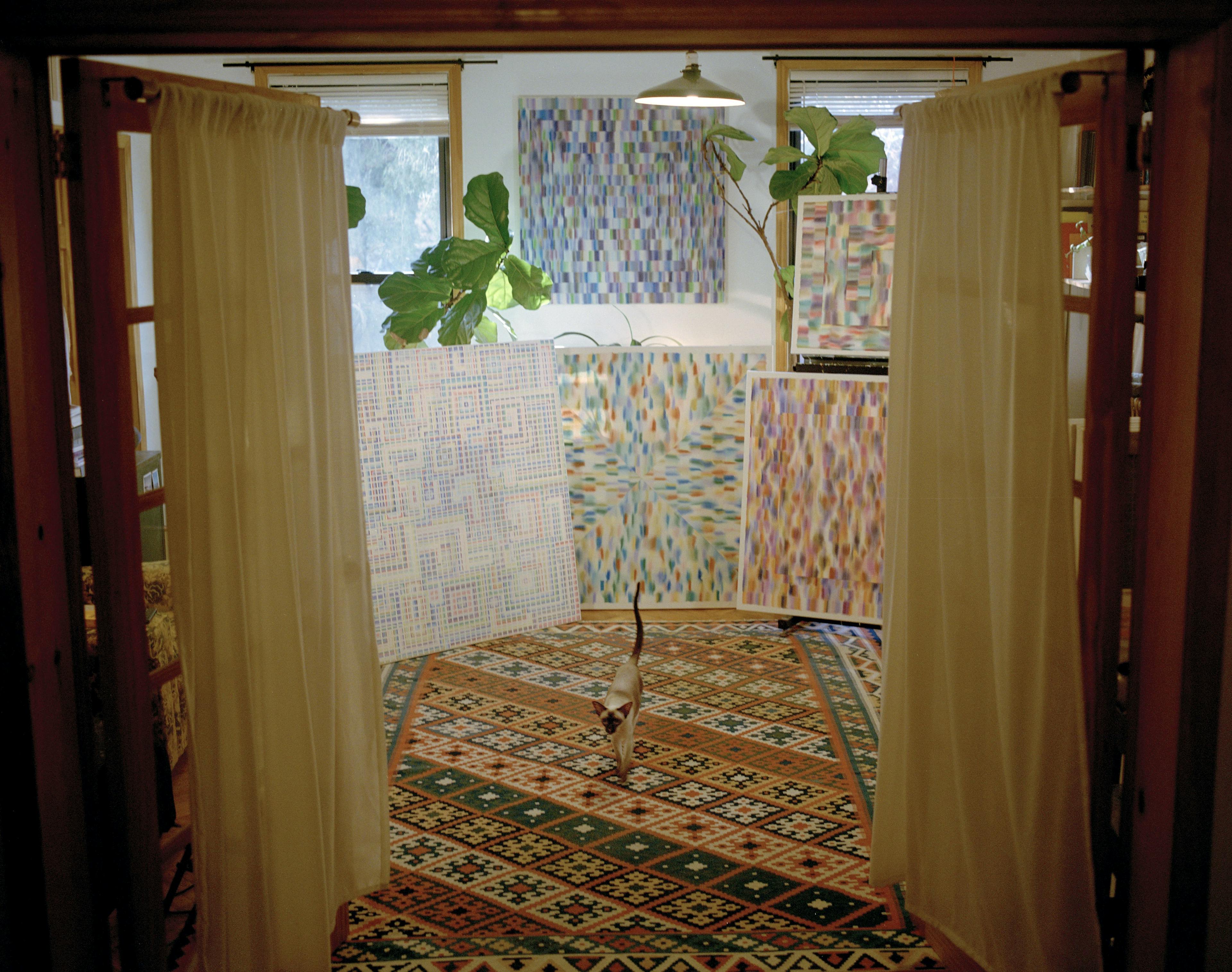Artist Devon Reina's studio with paintings, a geometric carpet, and a cat walking towards open double doors.
