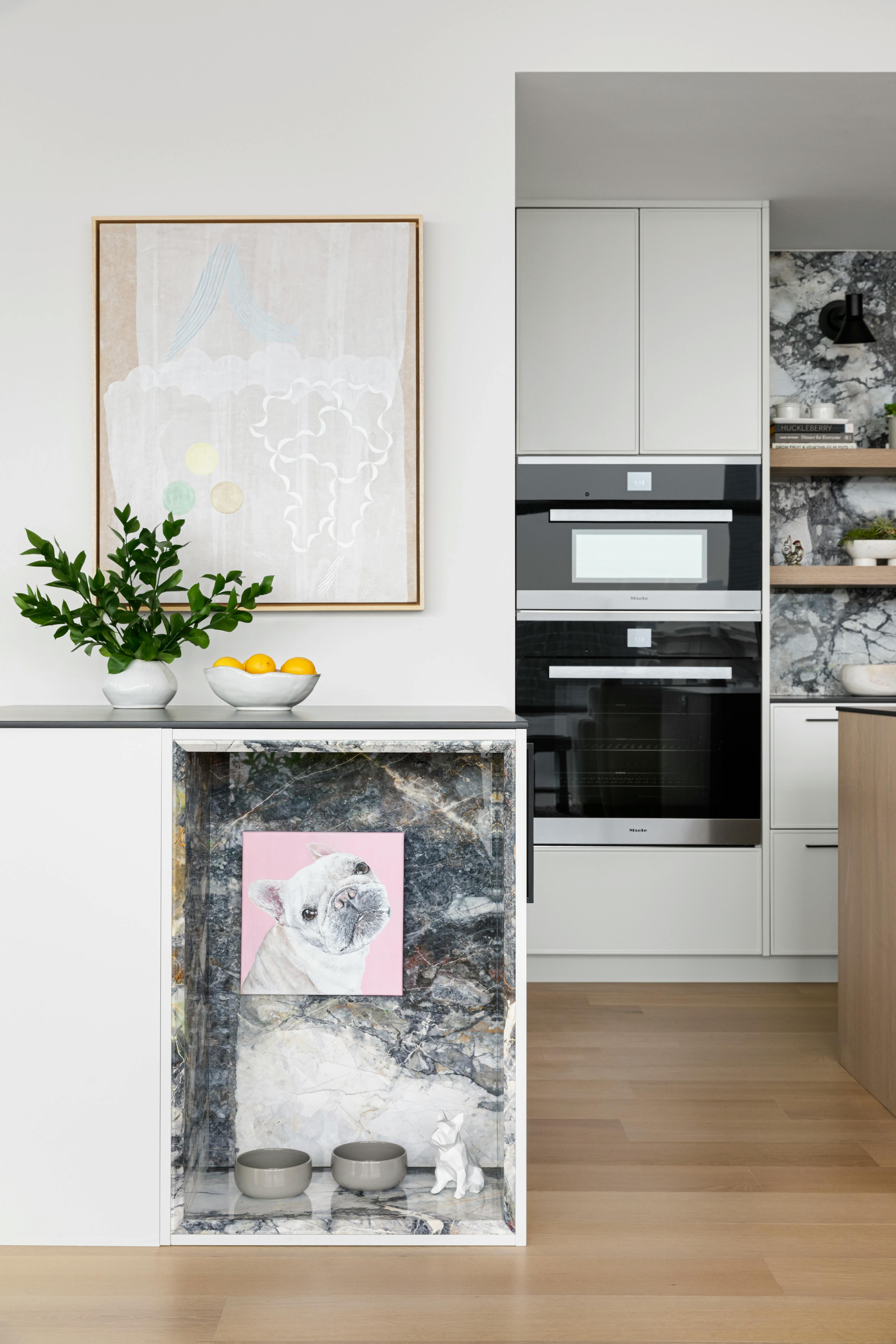 Abstract, neutral painting by artist Lydia Bassis installed in a modern kitchen above an open cabinet with a pink portrait of a french bull dog.