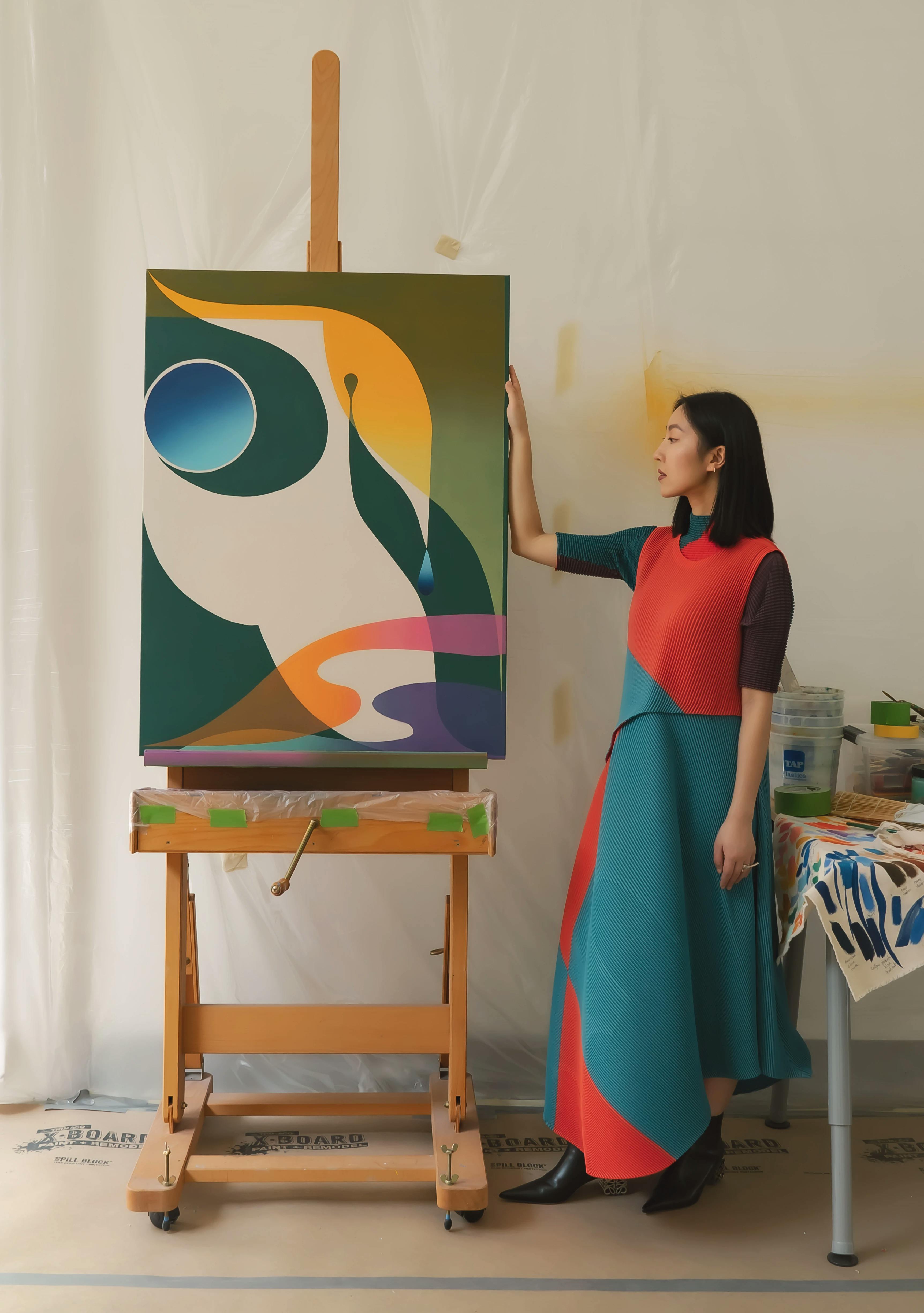 Artist Erin Zhao in blue and red dressing standing next to her painting, Letter To The Unknown #15, on an easel in her studio.