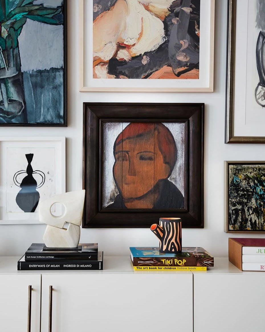 A gallery wall featuring a framed work of a black vessel by artist Vicki Sher above a white cabinet with a small abstract sculpture by artist Fitzhugh Karol displayed on top of it.