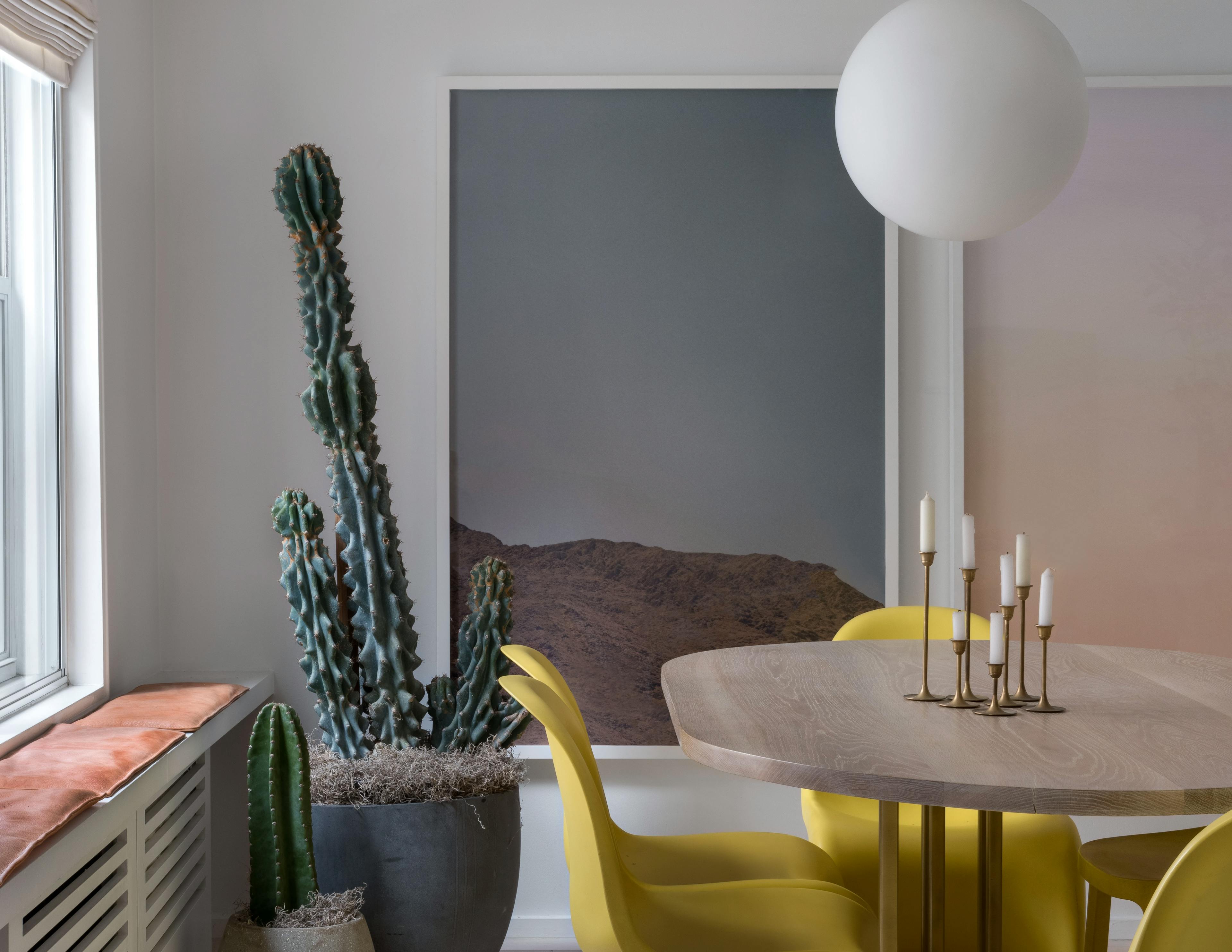 A large framed photograph of a mountain landscape by artist Jordan Sullivan on a white wall next to a cactus plant and behind a circular kitchen table with yellow chairs.