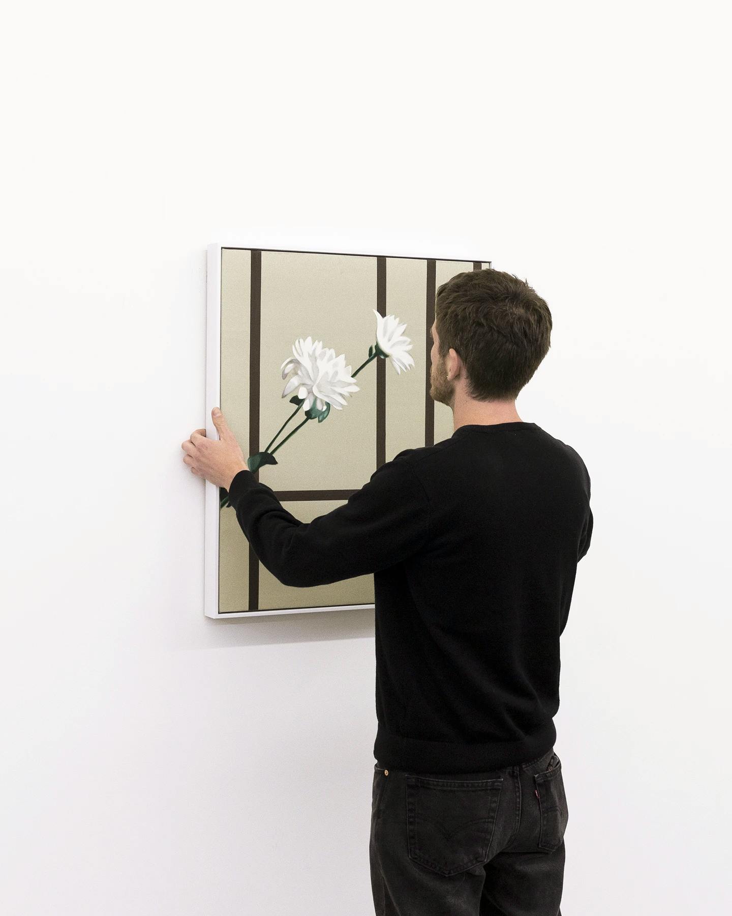 Artist Bryce Anderson wearing a black long-sleeve shirt and holding up his painting with a white flower against a white wall.