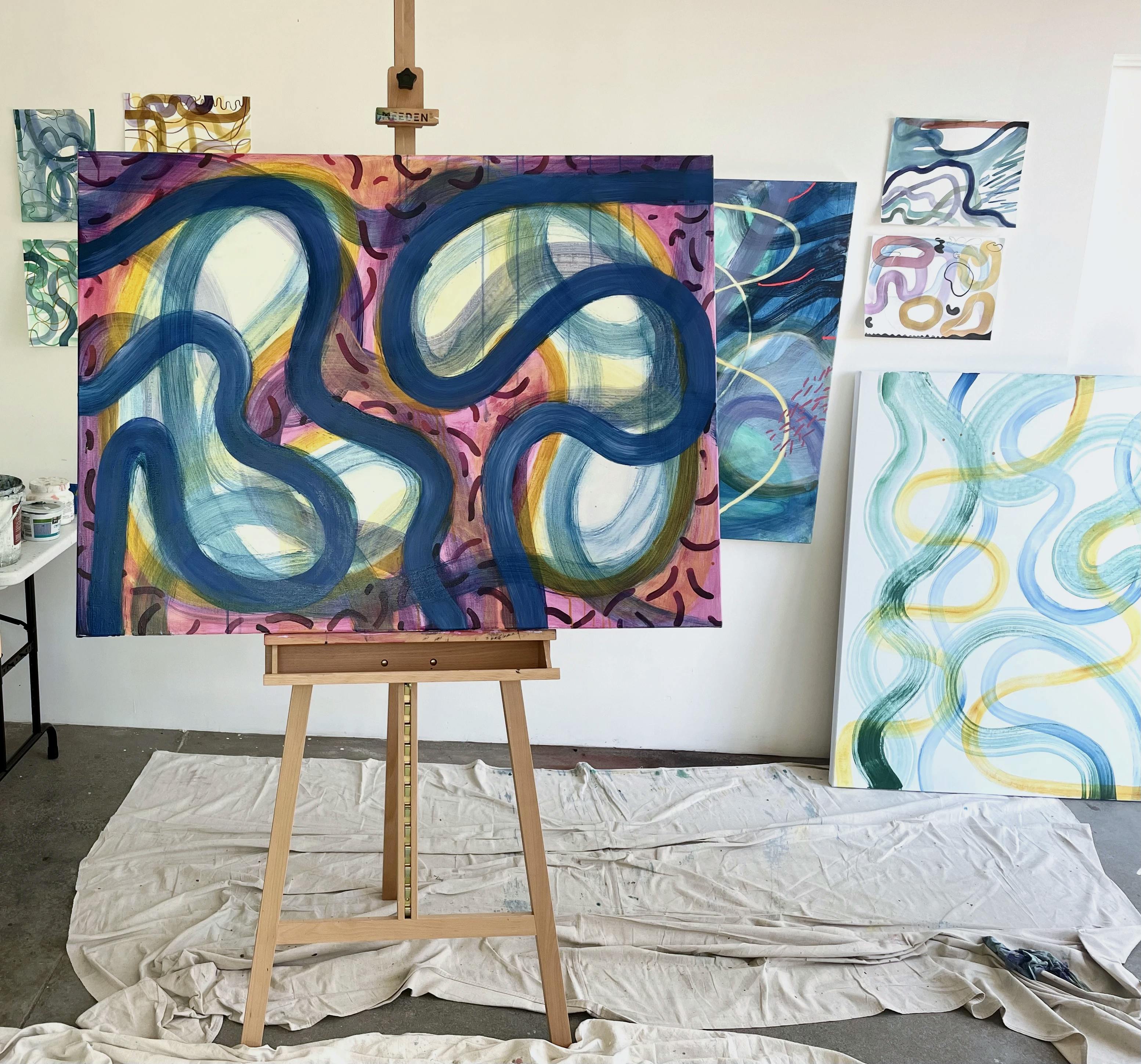 Abstract, gestural paintings of water landscapes, one on an easel and others mounted on a white wall, inside artist Aliza Cohen's studio.