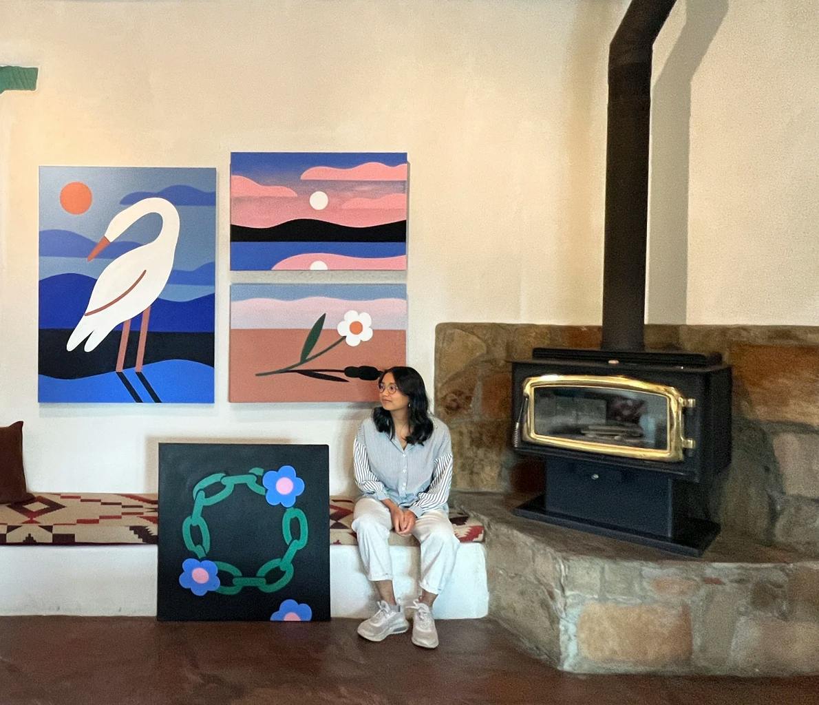 The artist Paulina Ho sits next to a fireplace surrounded by three of her large paintings depicting a crane, a sunset, and a daisy chain. 