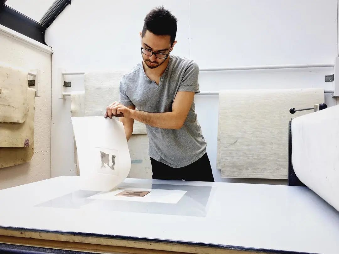 The artist Ruben Castillo standing in his studio pulling a print from the plate on his press. 