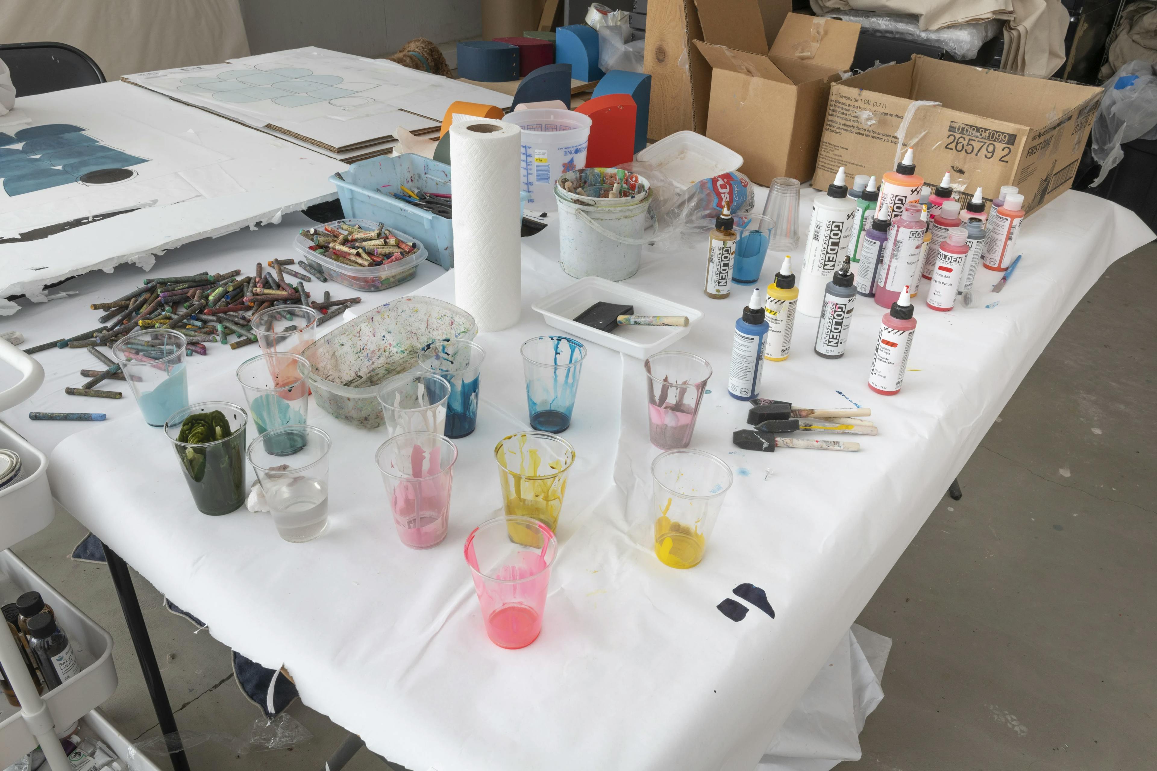 Piles of oil pastels and bottles of paint on a table in artist Vicki Sher's studio.