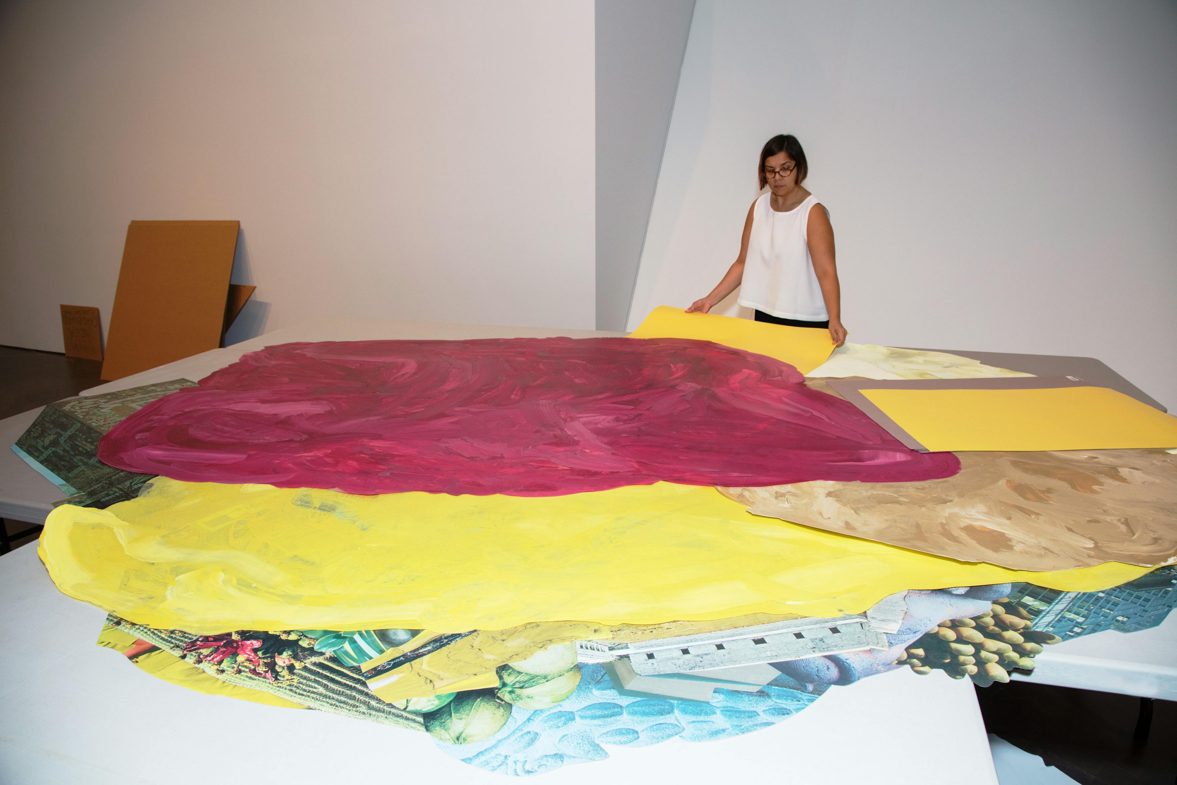 Artist Xochi Solis standing in front of table with a pile with oversized, naturally-dyed paper and printouts of collage material.
