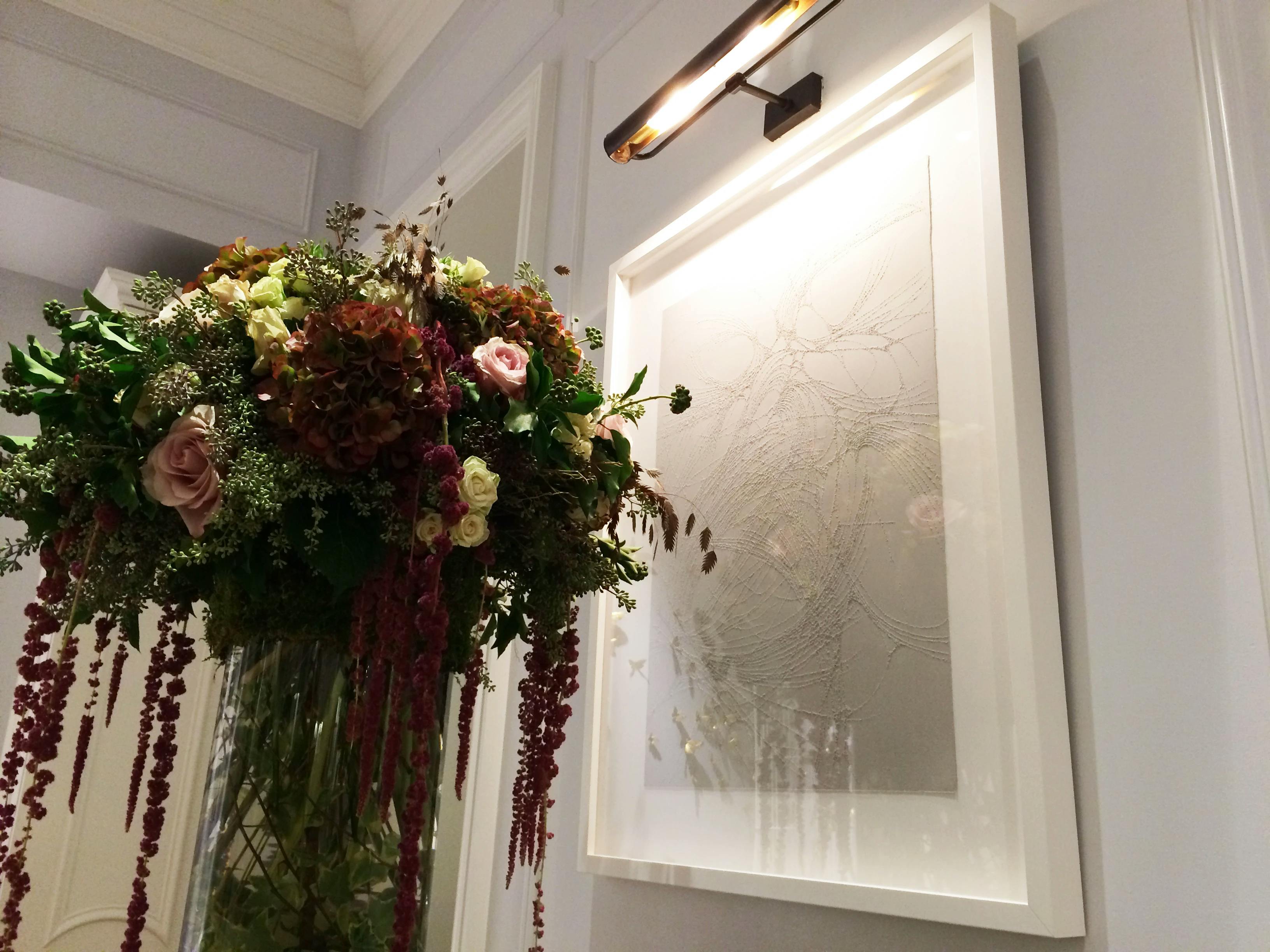 A framed artwork with carved, interlacing lines by artist Colleen Ho on a gray wall next to a large bouquet of cascading flowers.