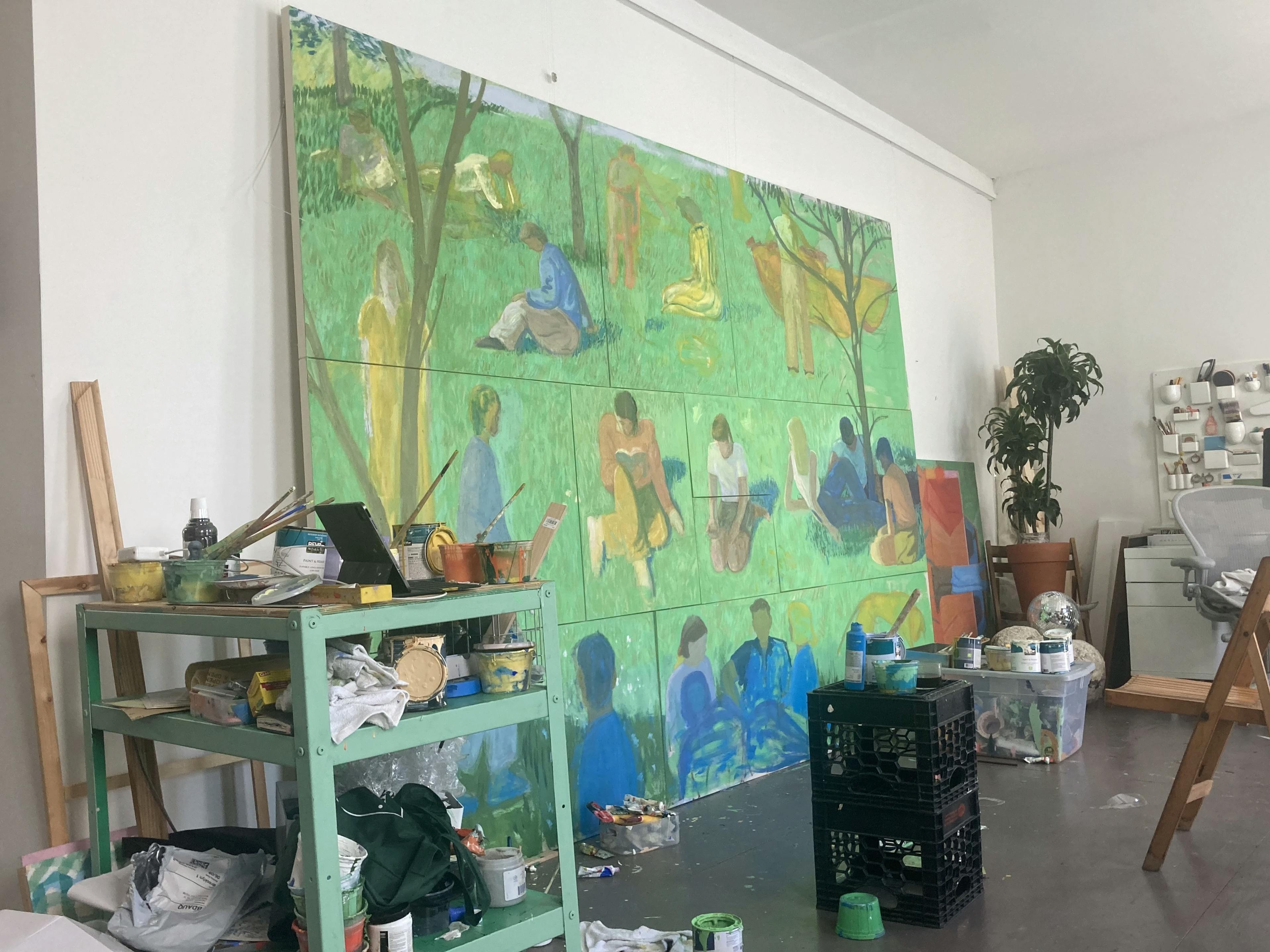 A large-scale installation comprised of multiple paintings of figures sitting in a grassy park by artist Jackson Joyce leaning against a white wall in his studio.