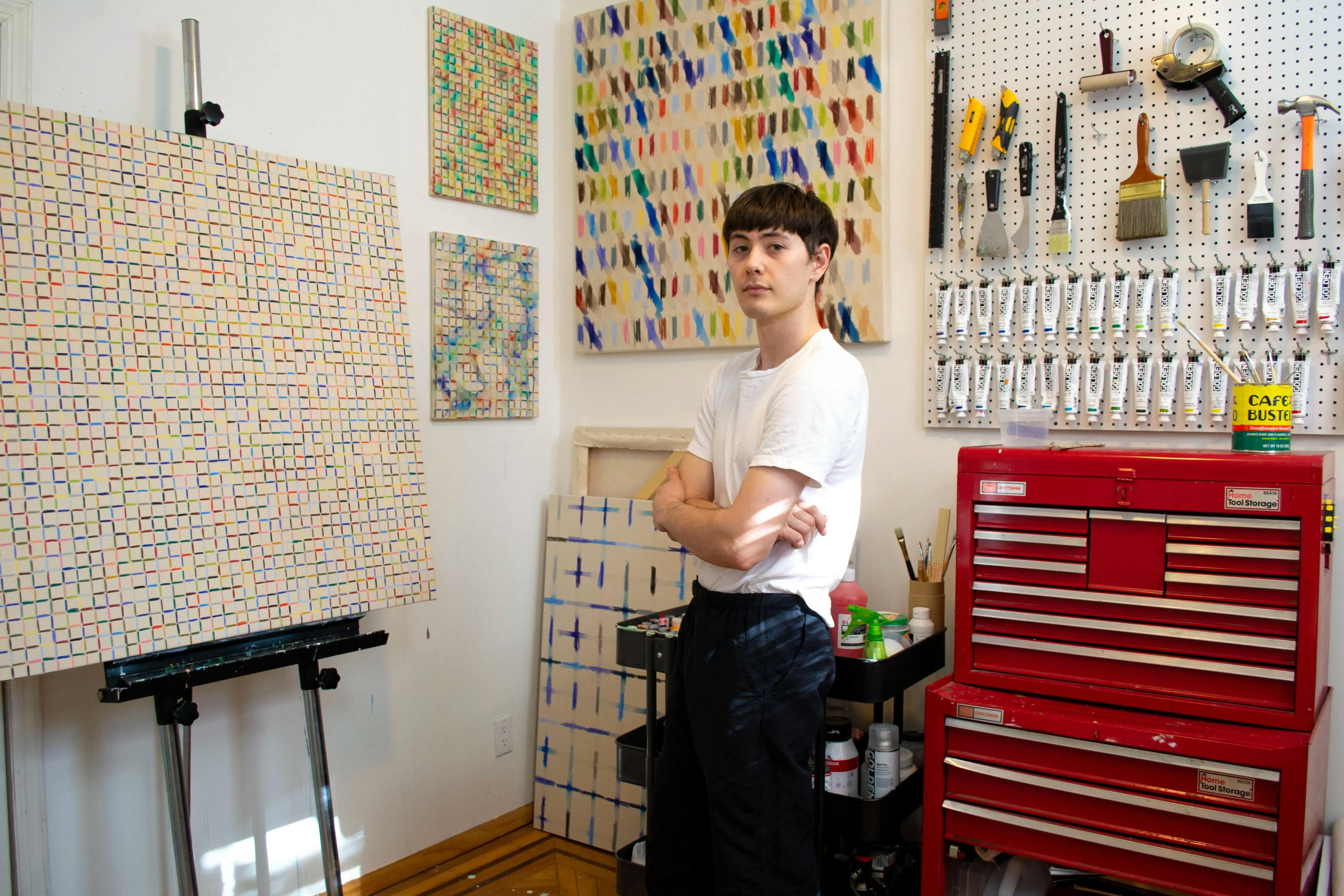 The artist Devon Reina standing in his studio surrounded by his paintings and neatly organized supplies. 