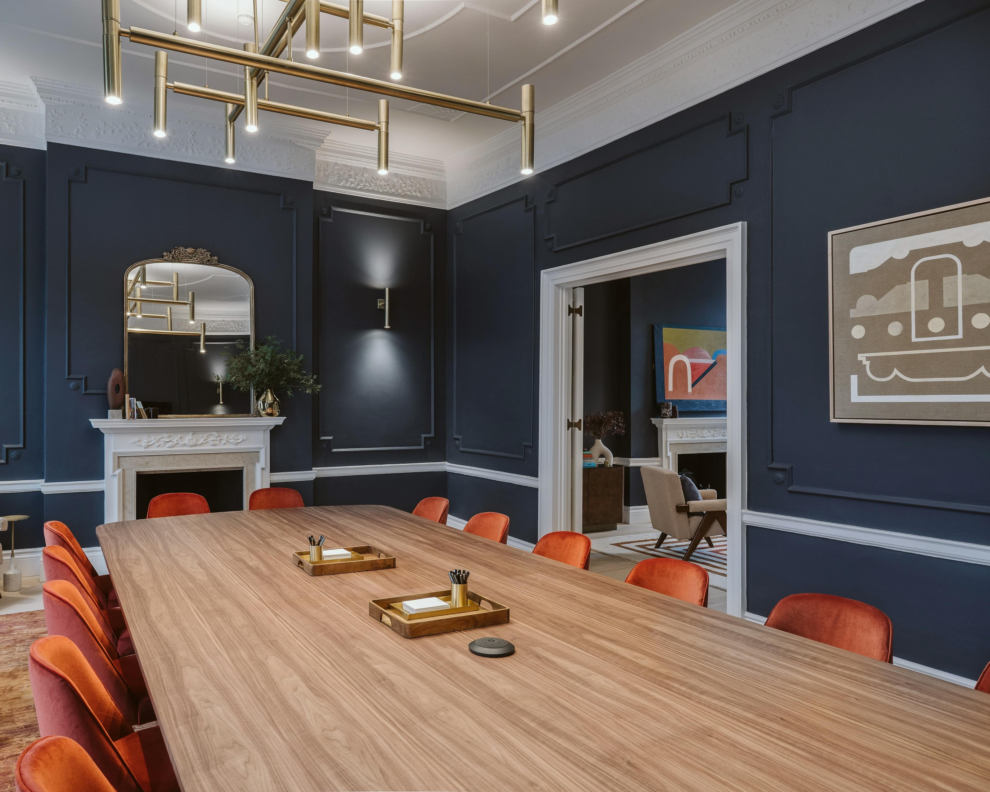 A conference room at Chief London with a long wooden table, orange chairs, and two original paintings, one by artist Kristin Texeira and one by artist Carla Weeks, installed on dark blue walls.