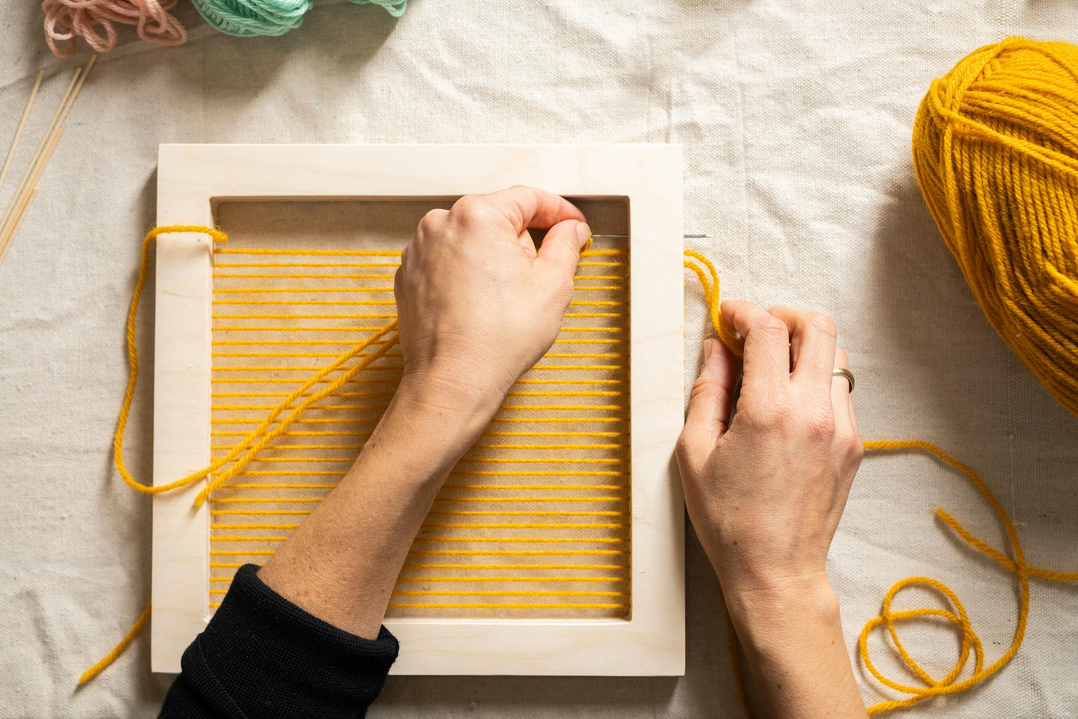 Artist Hayley Sheldon's hands pulling yellow yarn through a wood frame at MacArthur Place.
