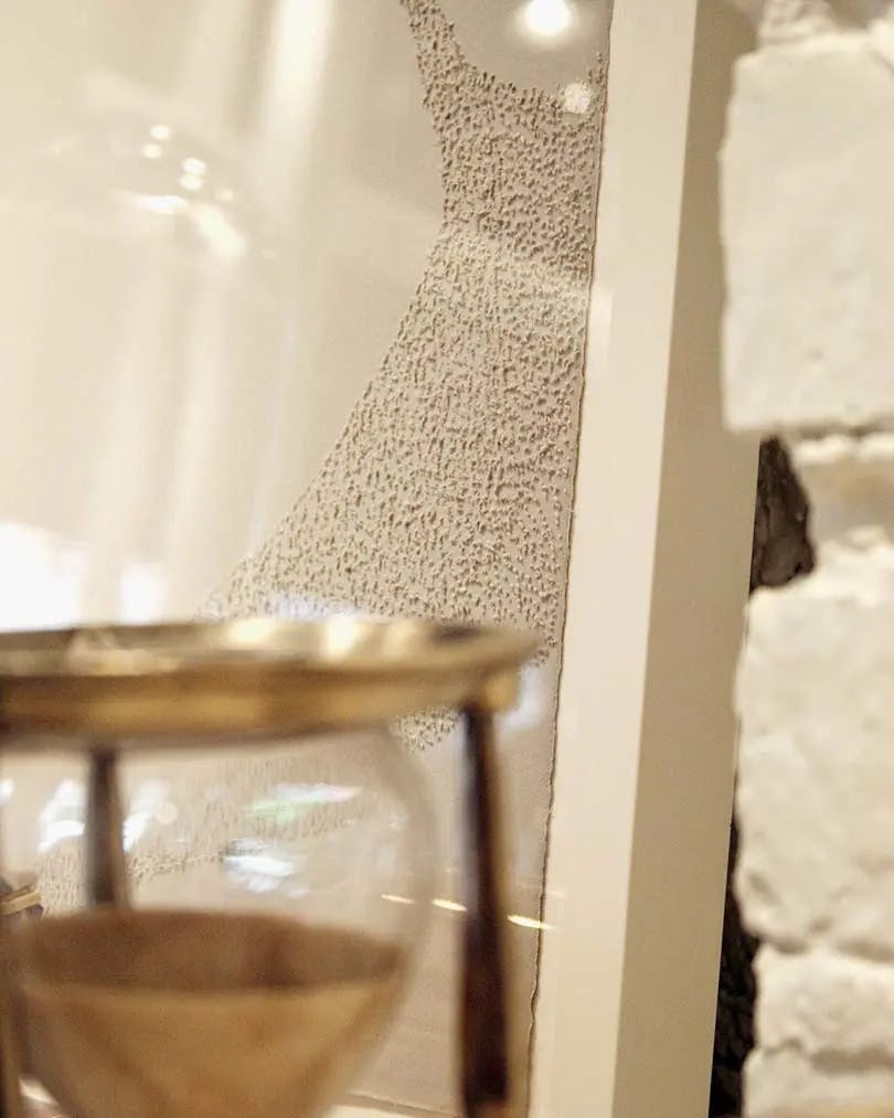 A close-up of a textured, neutral artwork by artist Colleen Ho behind a metal hourglass.