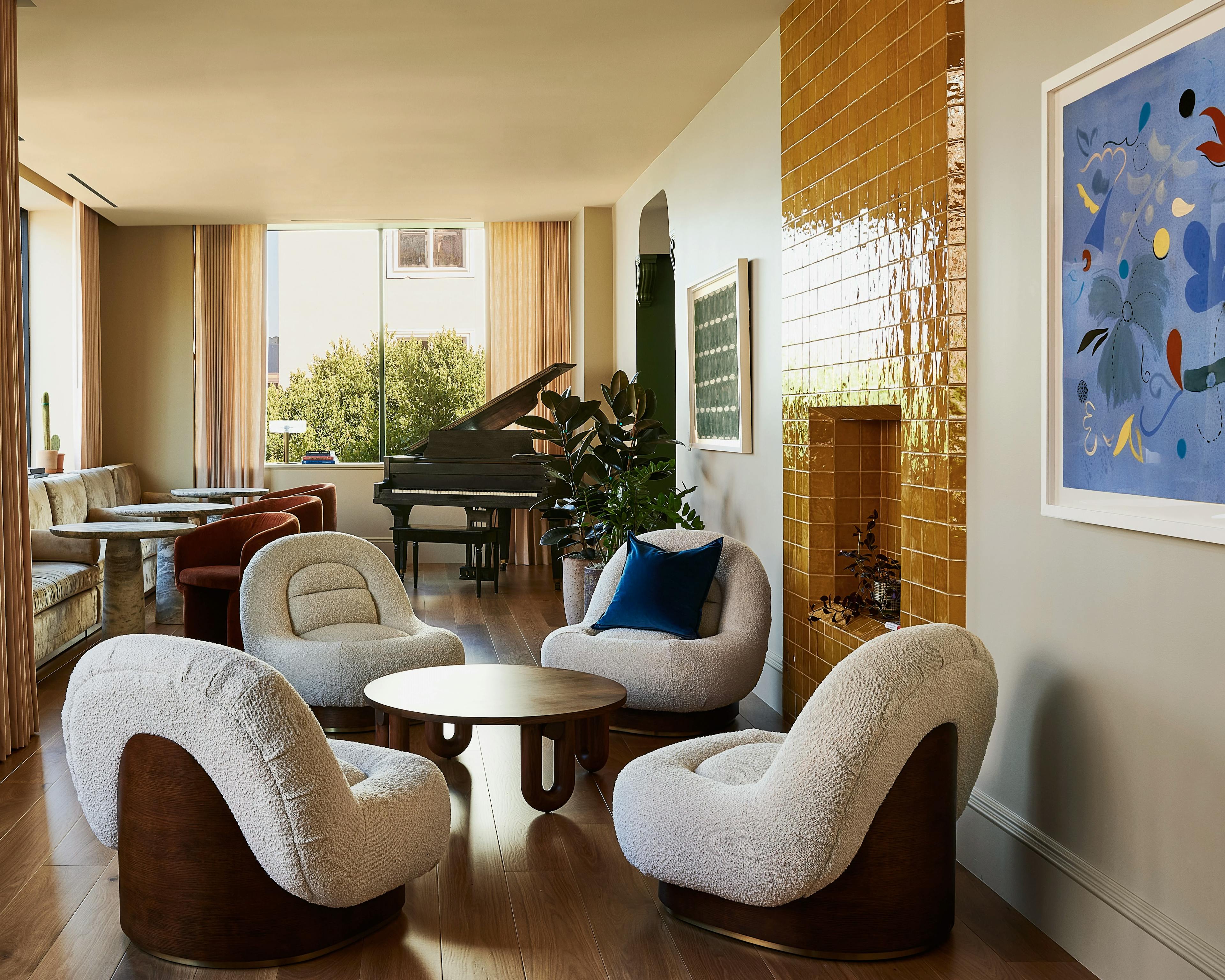 A blue floral painting by artist Kayla Plosz Antiel and geometric artwork by artist Katrine Hildebrandt-Hussey installed in a lounge area at the Chief San Francisco Clubhouse with four arm chairs and a baby grand piano in the back.