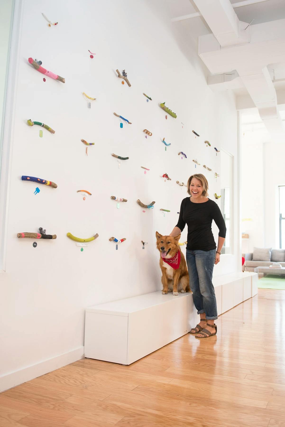 Artist Christina Watka standing next to a dog with a red bandana alongside a white wall with an installation of fourty-three colorful stick sculptures.