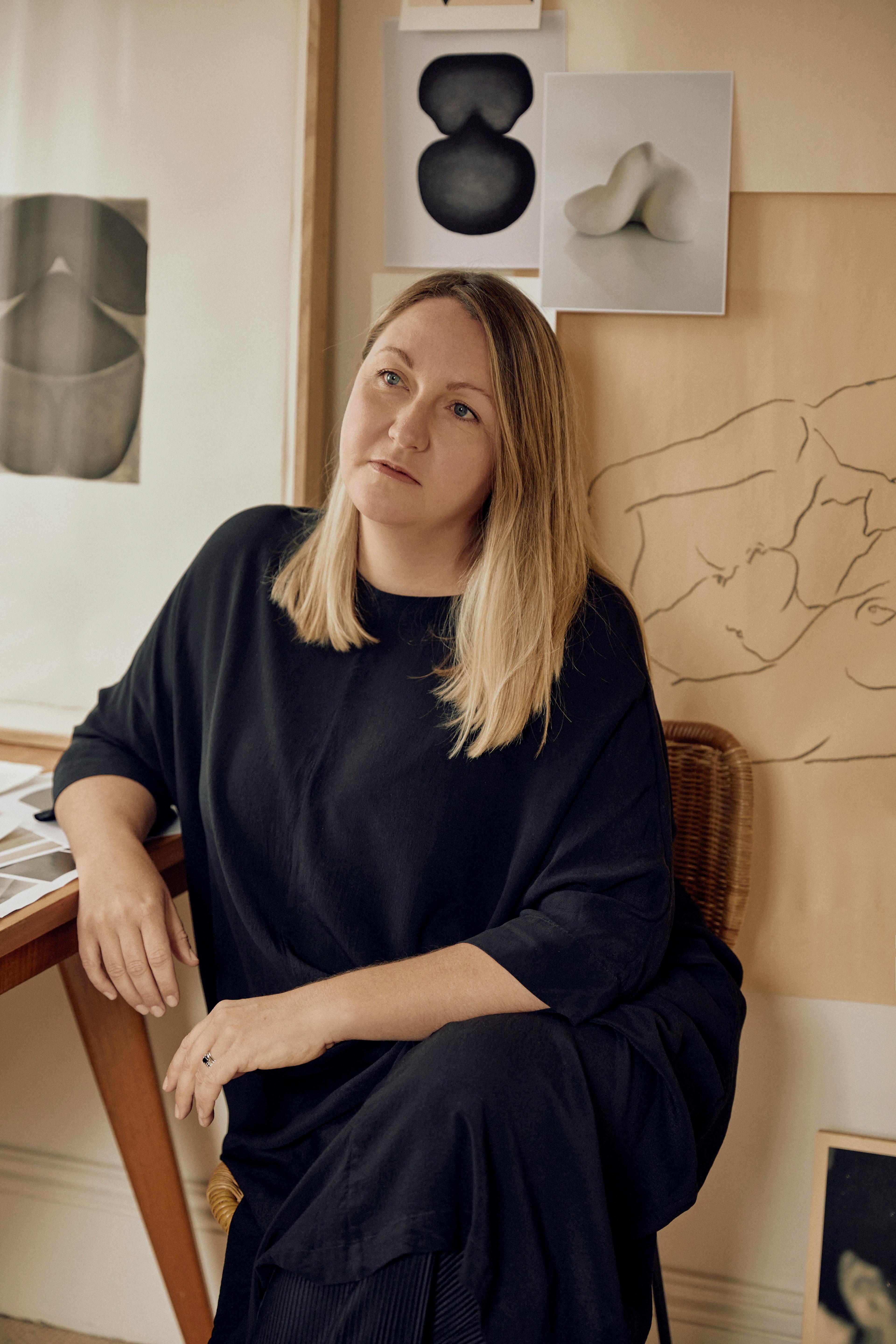 Artist Caroline Walls, dressed in all black, sits on a chair at her studio and looks off into the distance.