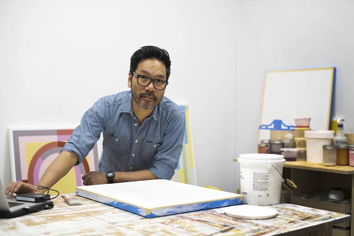 Artist Christian Nguyen wearing a denim shirt, standing in his studio surrounded by paintings on canvas.