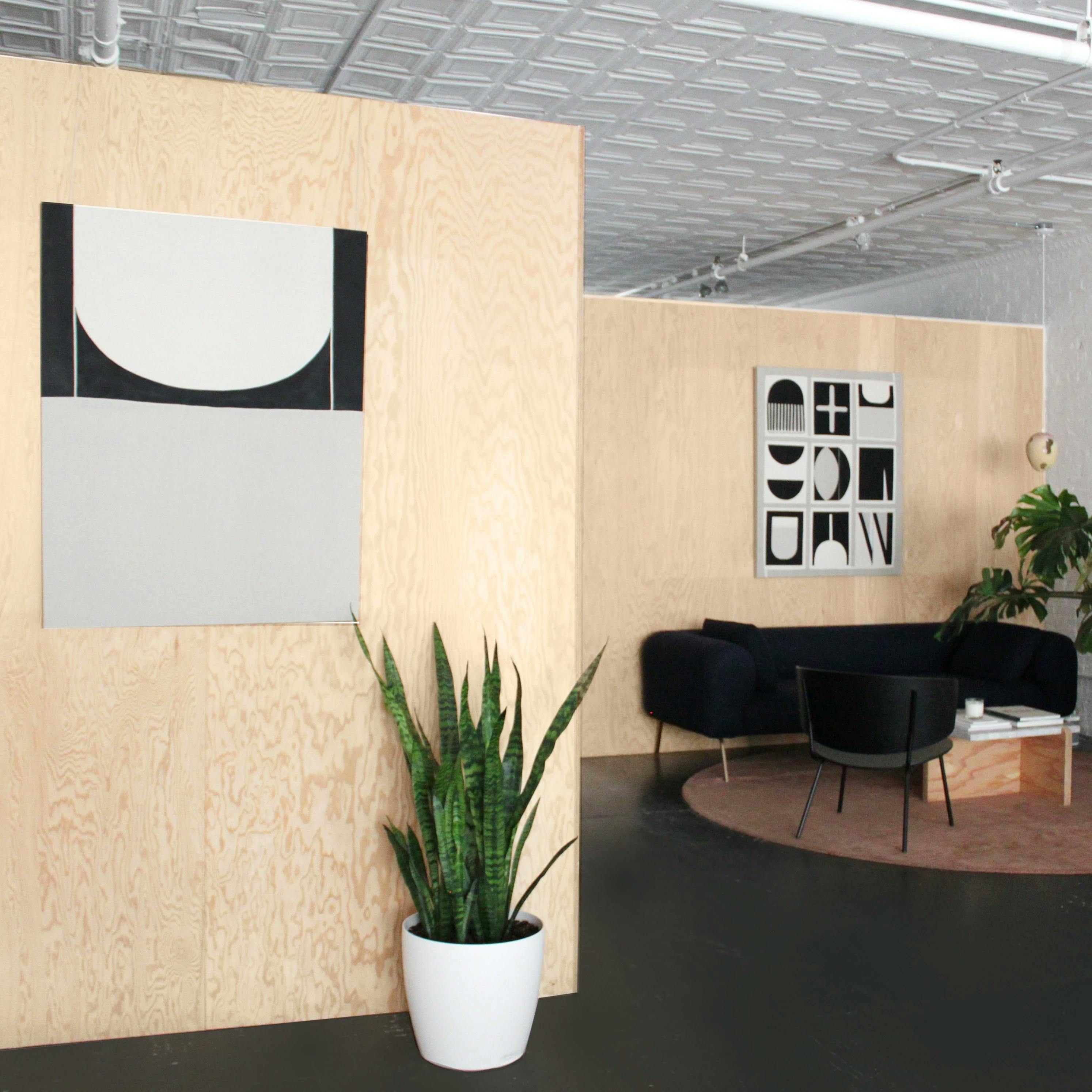 Two gray, white, and black geometric paintings by artist Hyun Jung Ahn, one on a wood wall above a green potted plant and the other above a blue sofa, on display at the Uprise Art showroom. 