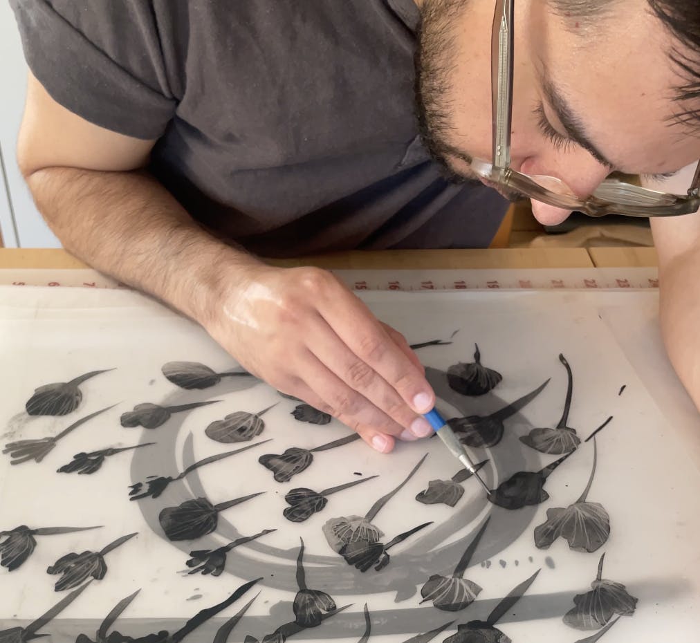 Aerial view of artist Ruben Castillo using a thin ink pen to draw repeating floral motifs on translucent paper.