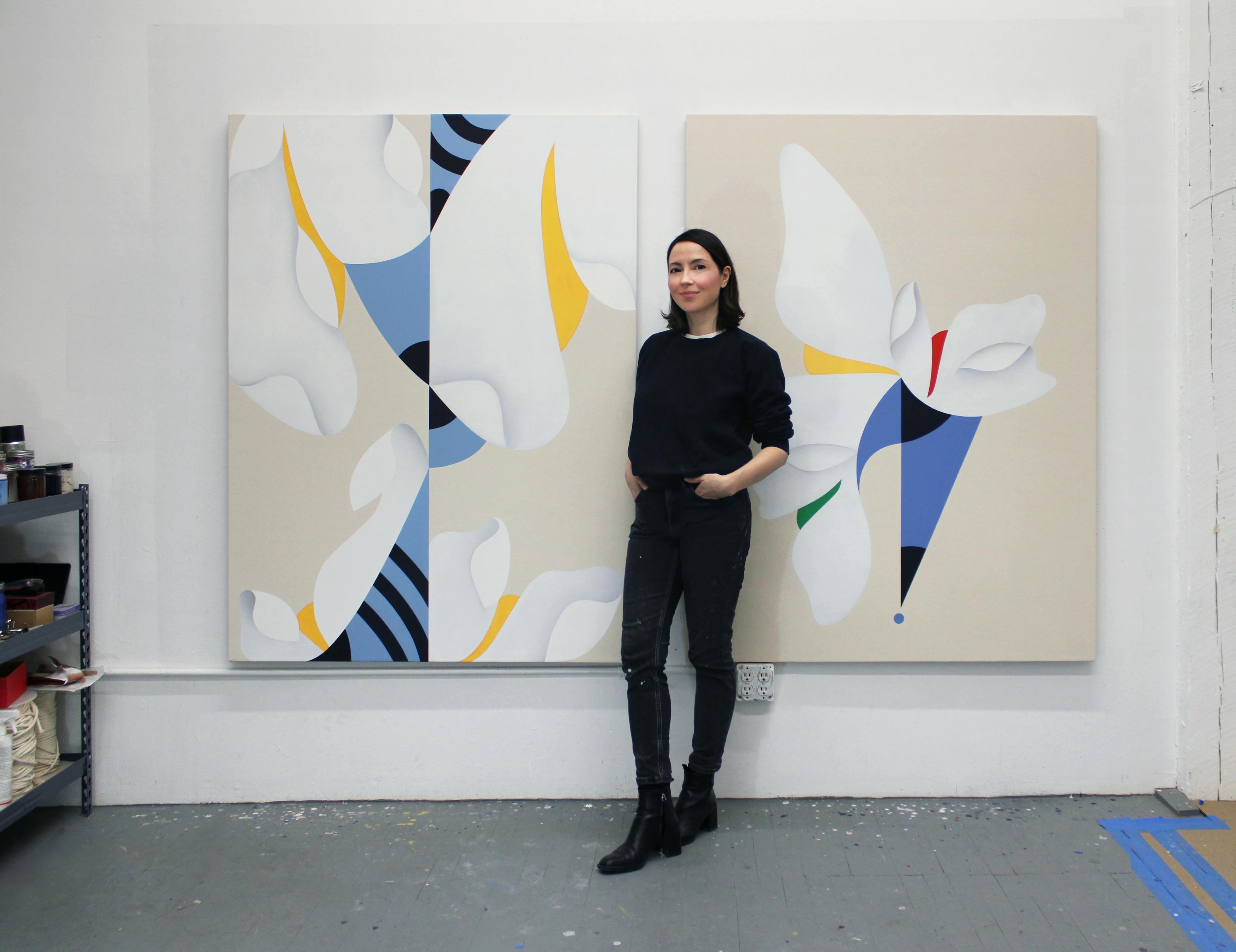 Artist Senem Oezdogan standing in front of two large blue, yellow and white paintings in her studio. 