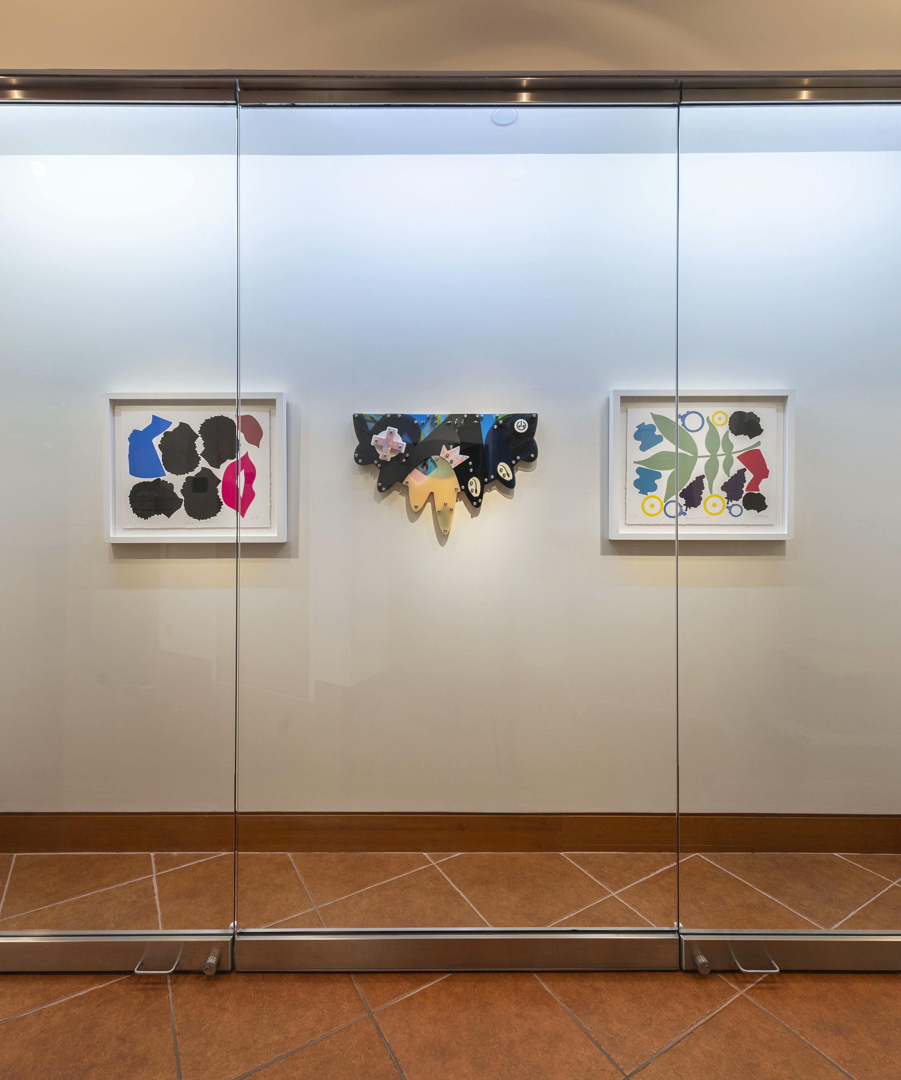Three works by artist Damien Davis, two framed figurative prints and one black abstract wall sculpture, installed on a white wall behind a glass case at Montclair State University.