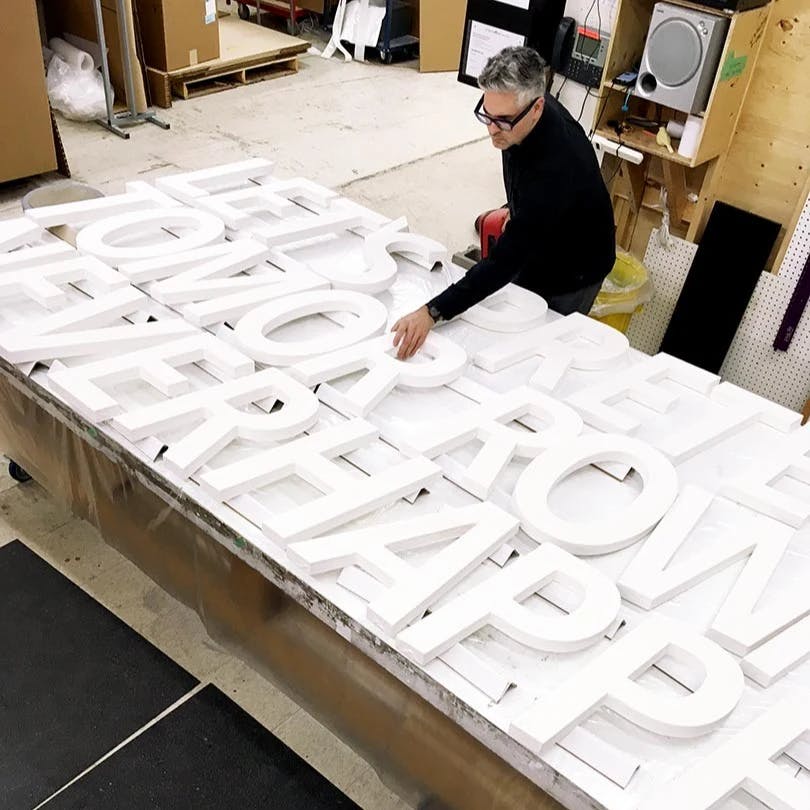 Artist Ben Skinner placing a large, white "R" within a line of sculptural letters that spells out "Let's Pretend Tomorrow Never Happens" on a table inside his studio.