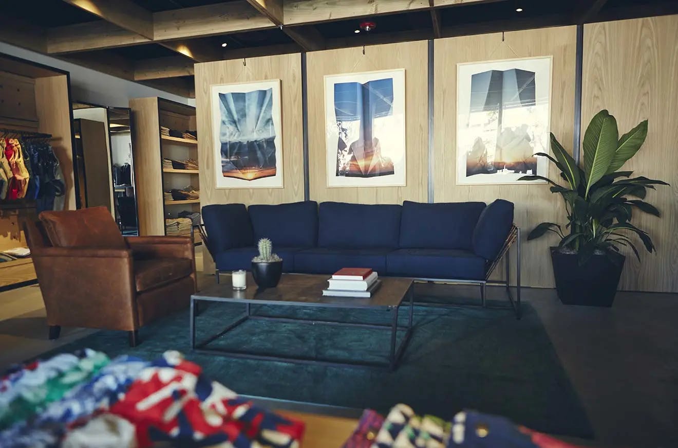 Three framed photographs of cloudy skies by artist Millee Tibbs on a wood wall above a blue sofa at a Bonobos store in Los Angeles.