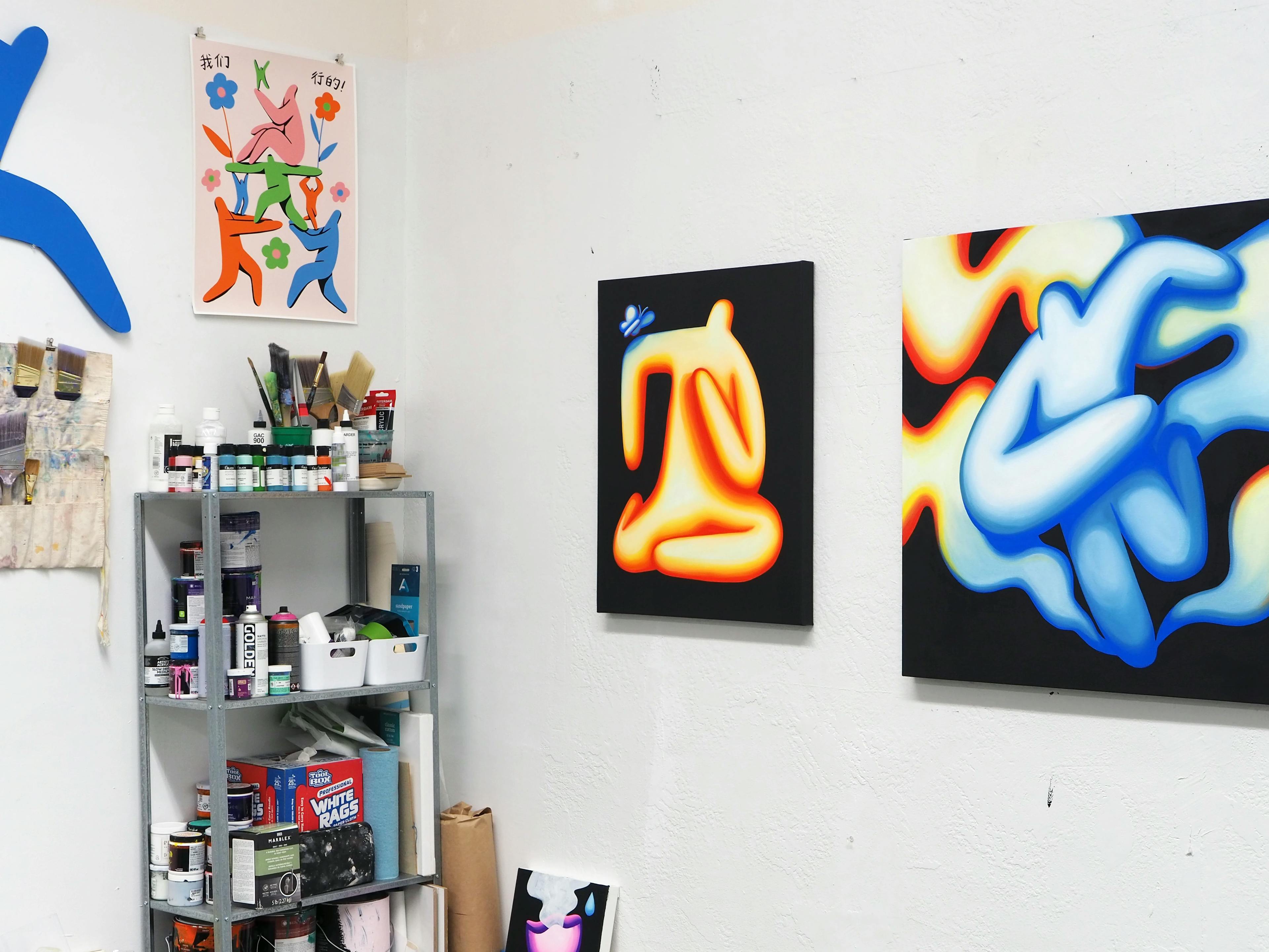 A corner view of Jocelyn Tsaih's studio. There is a standing shelf filled with supplies in the corner, and on the walls are three paintings of abstracted figures. 